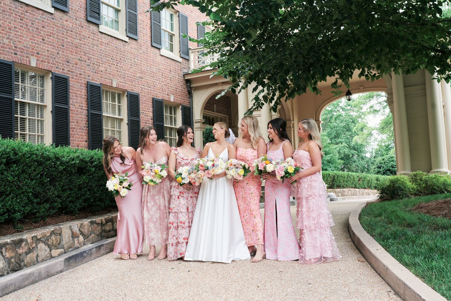 Bridesmaids in pink gowns gather with bride on curved walkway by brick building.