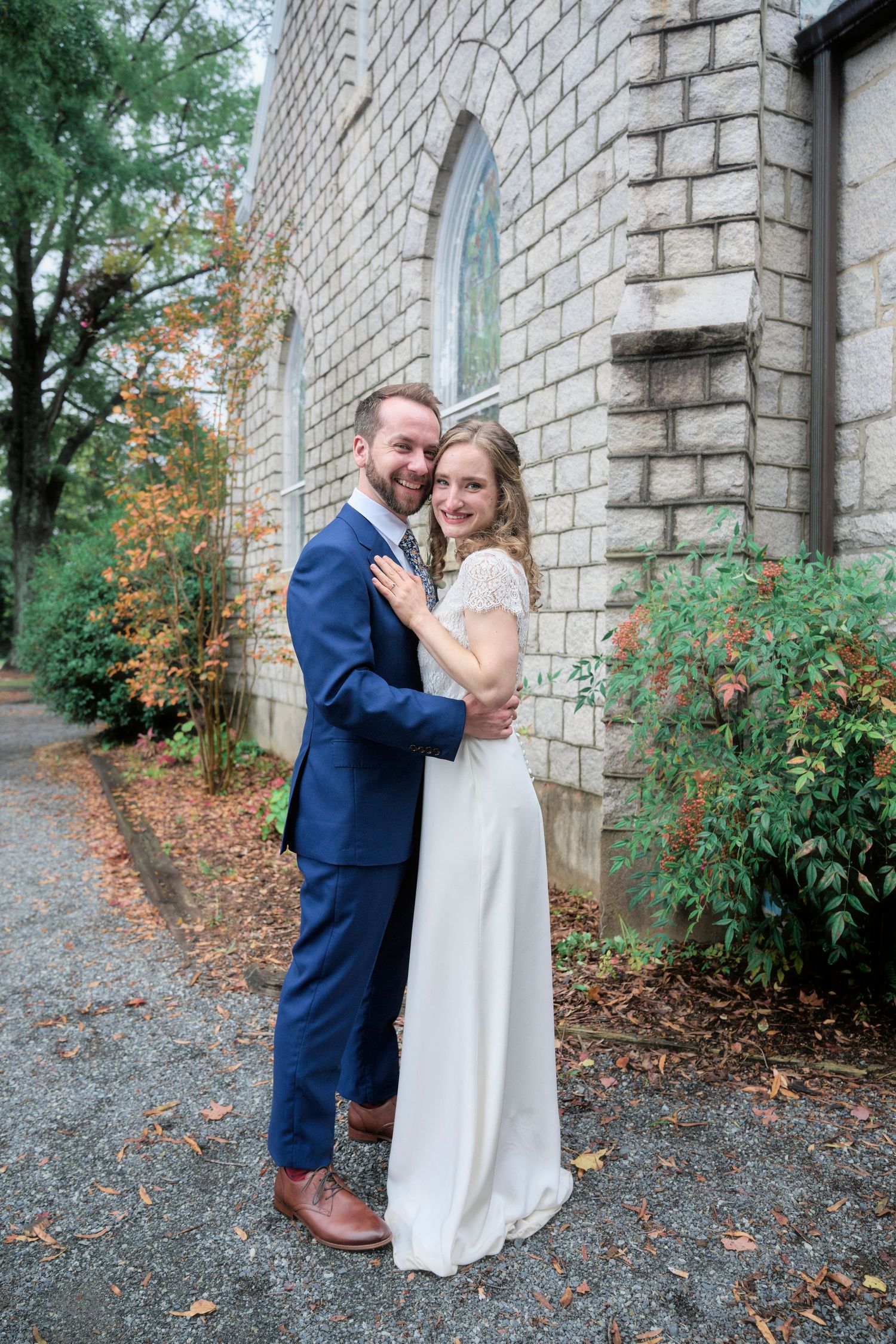 A full-length portrait of a couple in wedding attire embracing by a stone church with fall foliage.