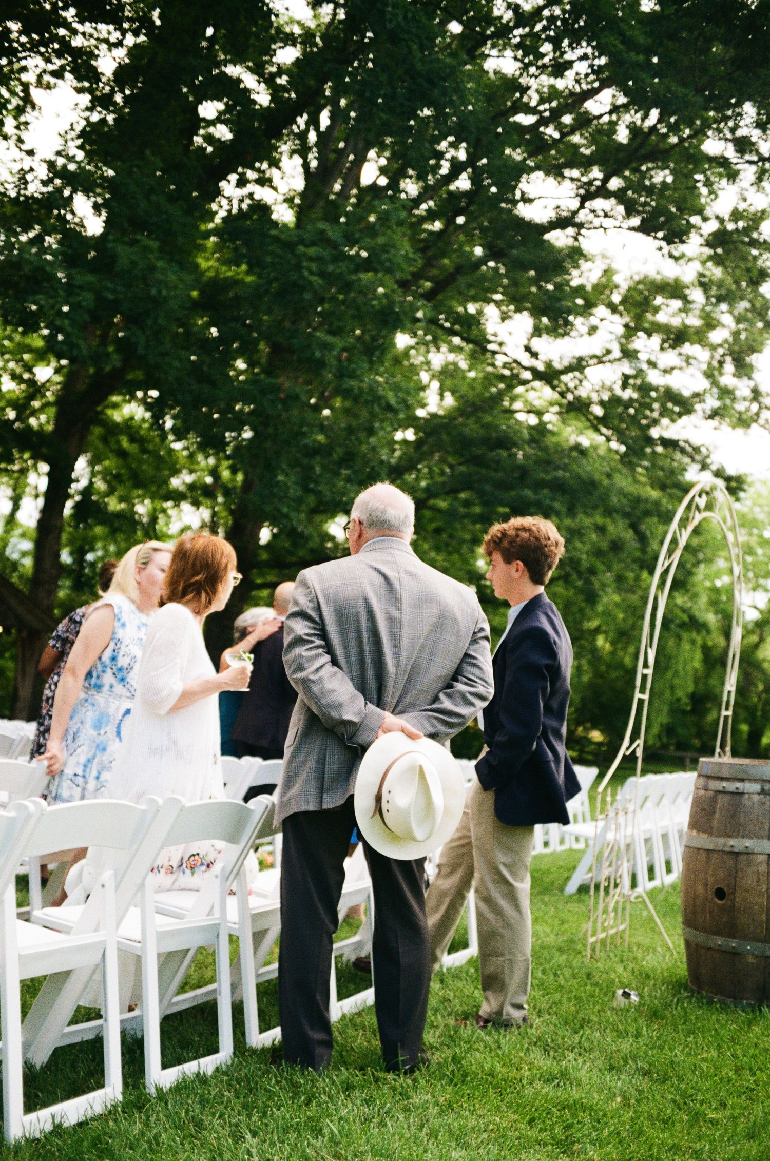 Wedding ceremony setup outdoors with white chairs and wooden barrels under shady trees.