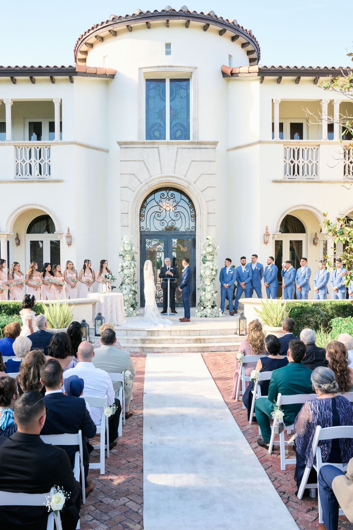 Wedding ceremony setup at Mediterranean villa with white aisle runner and blue-clad wedding party.