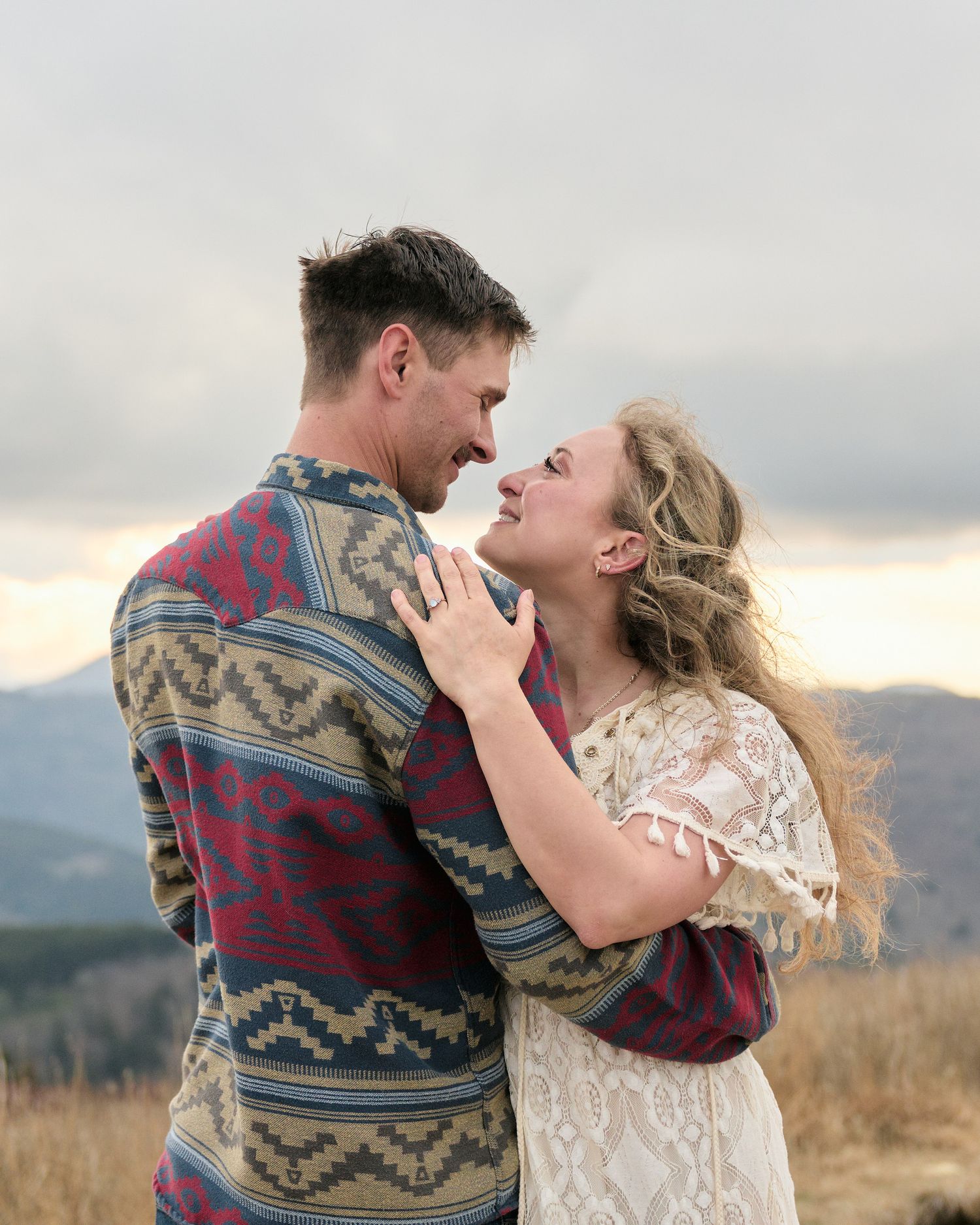 A couple shares a romantic kiss in a field at sunset while wearing southwestern style clothing and a lace dress.