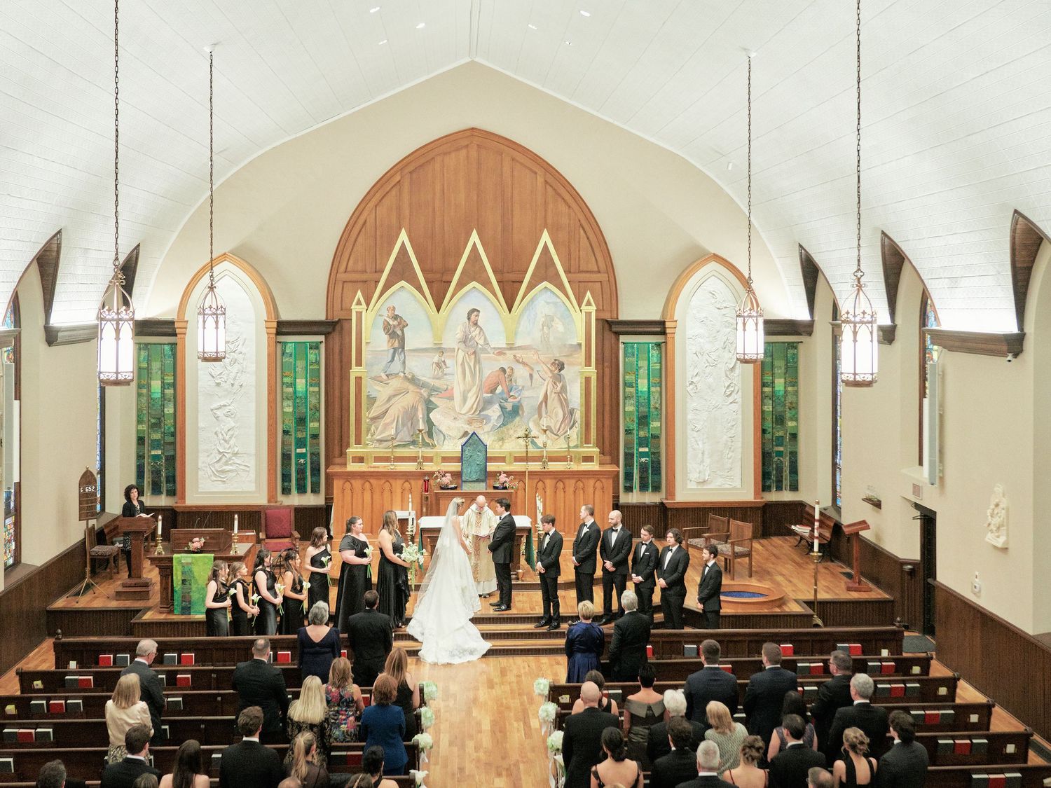 Wedding ceremony in a beautiful church with bride, groom, and wedding party at the altar, surrounded by stained glass windows.