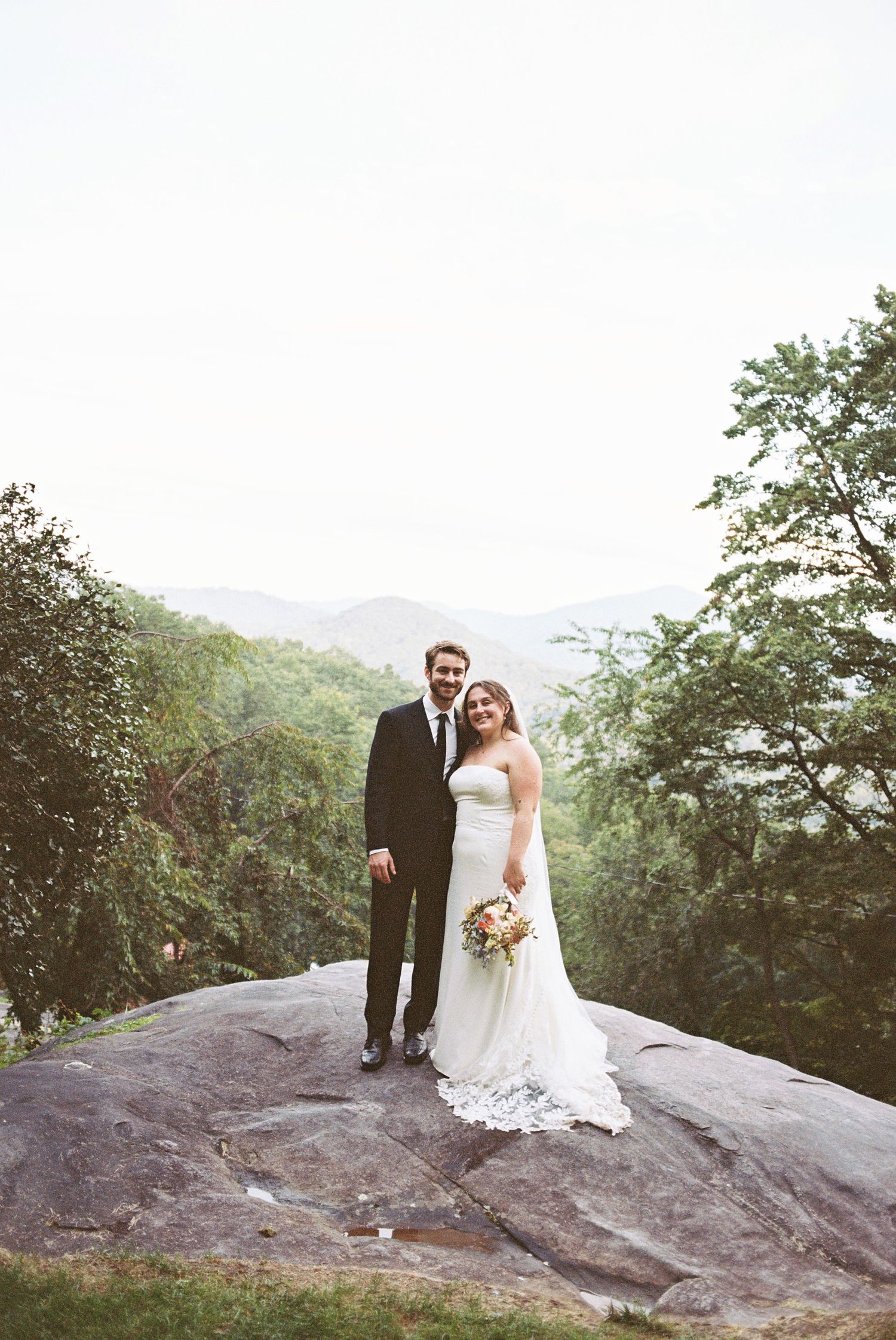 Bride and groom pose on a large rock with mountain views and lush green trees in the background.