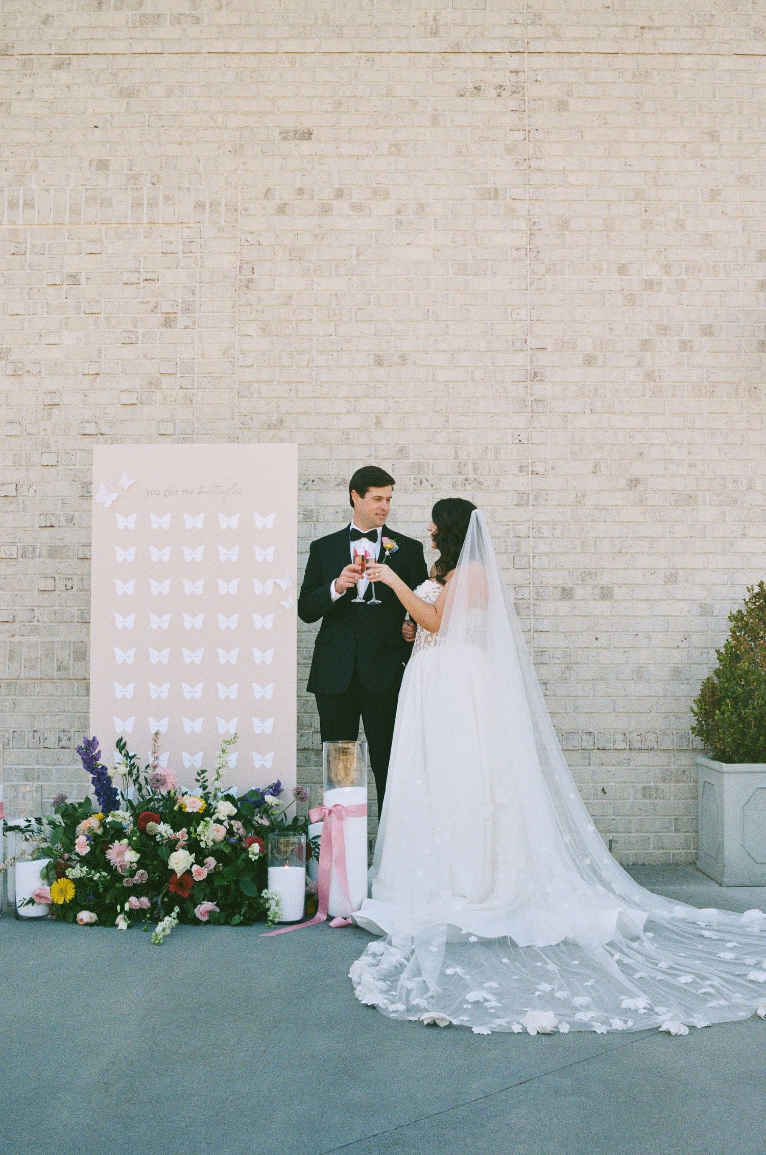 A wedding couple poses against a white brick wall with floral arrangements and decorative elements in a modern setting.