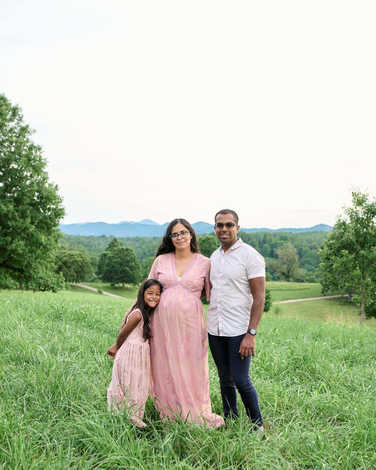 Pregnant woman in pink maxi dress poses with husband and daughter in lush green meadow with mountain views.