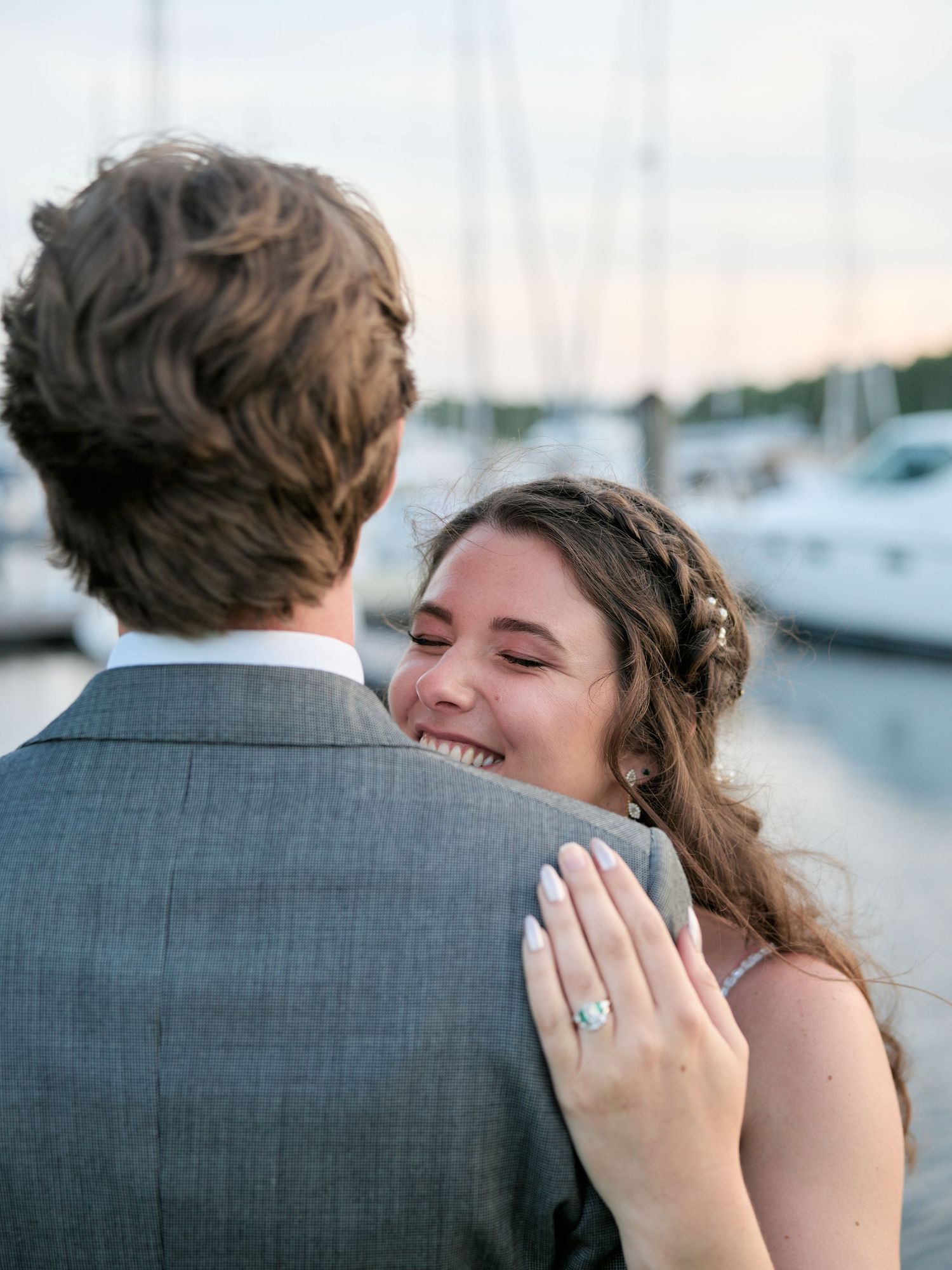 A joyful bride displays her engagement ring while embracing her groom near a marina at sunset.