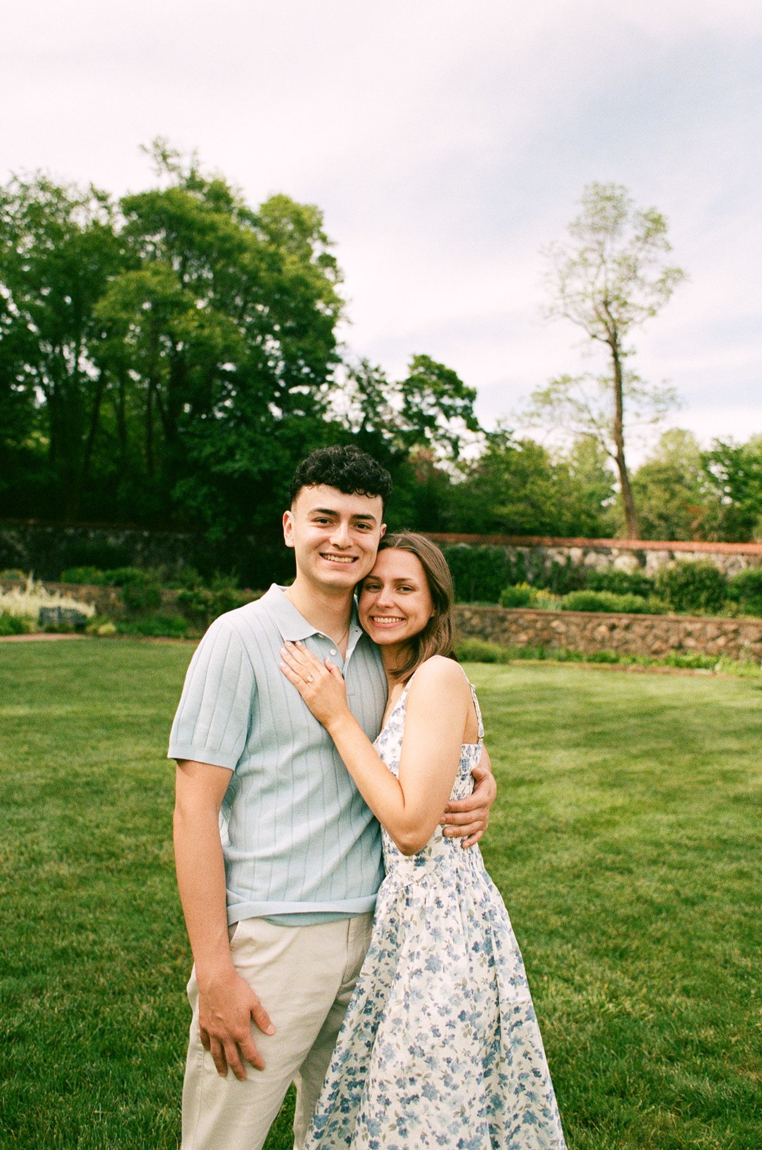 A smiling couple poses on a lush green lawn, the woman showing an engagement ring while embracing her partner outdoors.