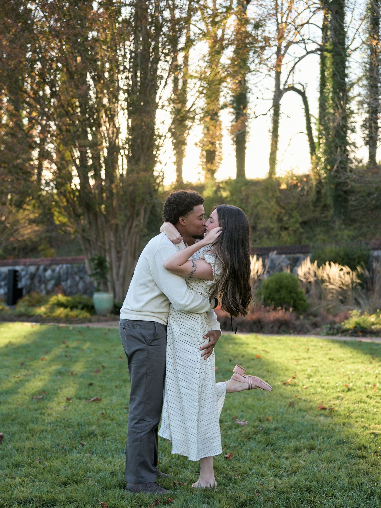 A couple shares romantic moments in a sunlit garden setting with trees in the background at sunset.