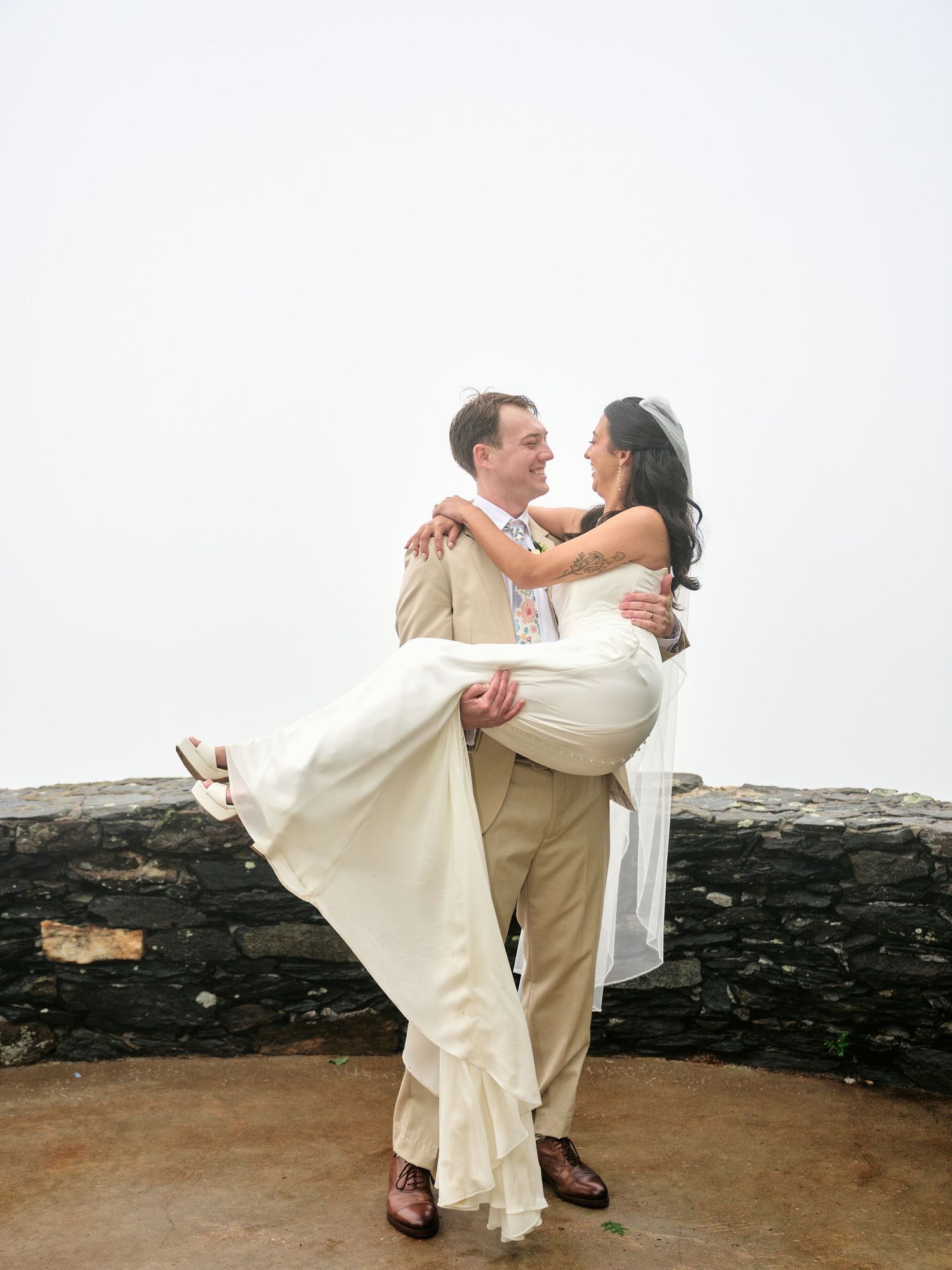 Groom lifts bride in white wedding dress outdoors against stone wall backdrop on their wedding day.
