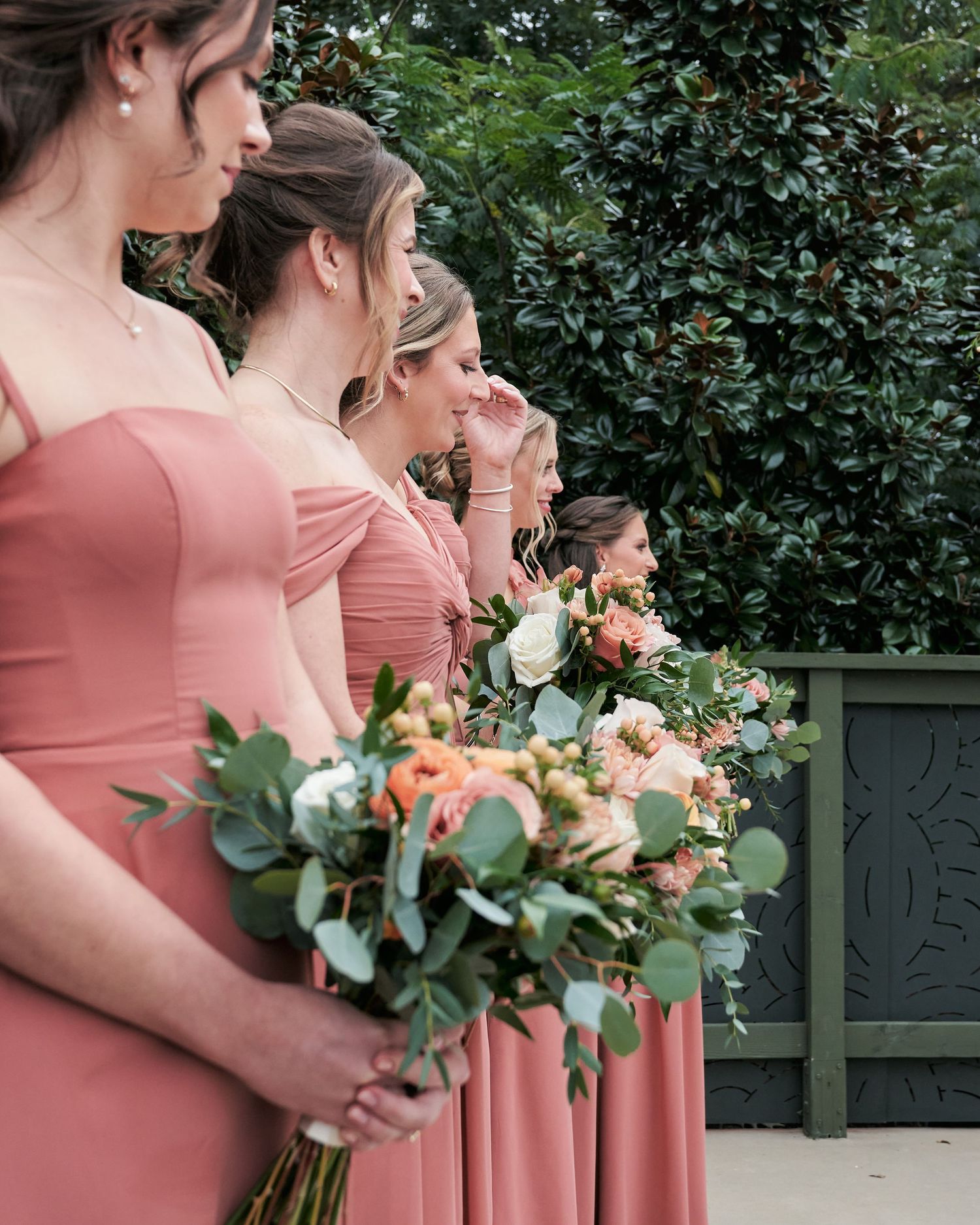 Bridesmaids in dusty rose off-shoulder gowns holding bouquets of peach and white flowers with eucalyptus greenery.