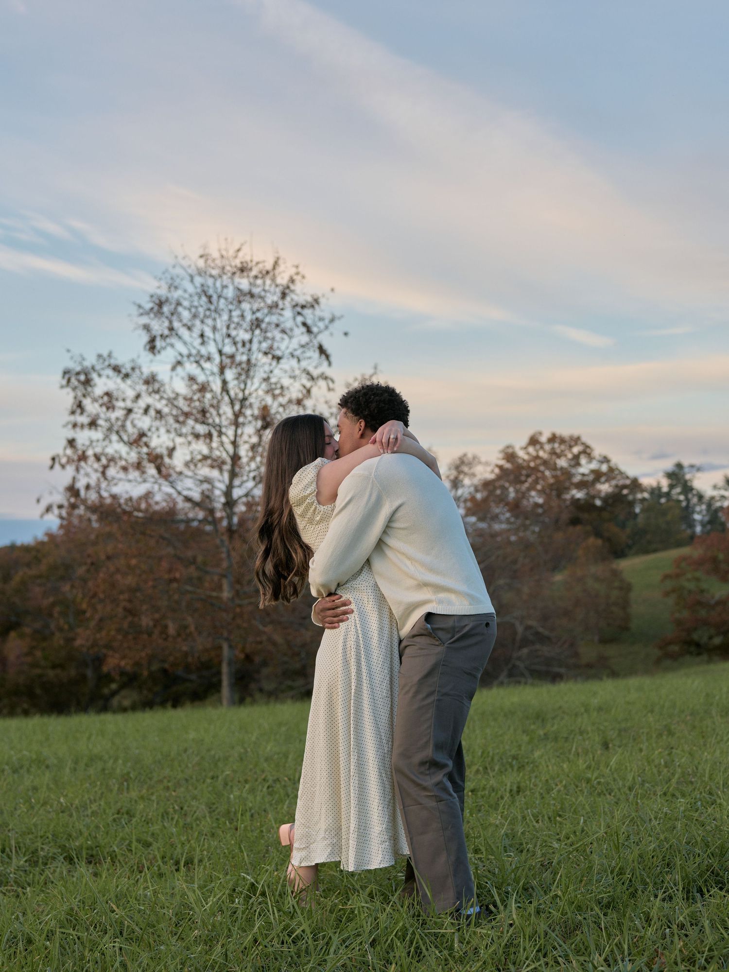 A couple shares a tender moment together in a grassy field during sunset, creating a romantic silhouette against the sky.