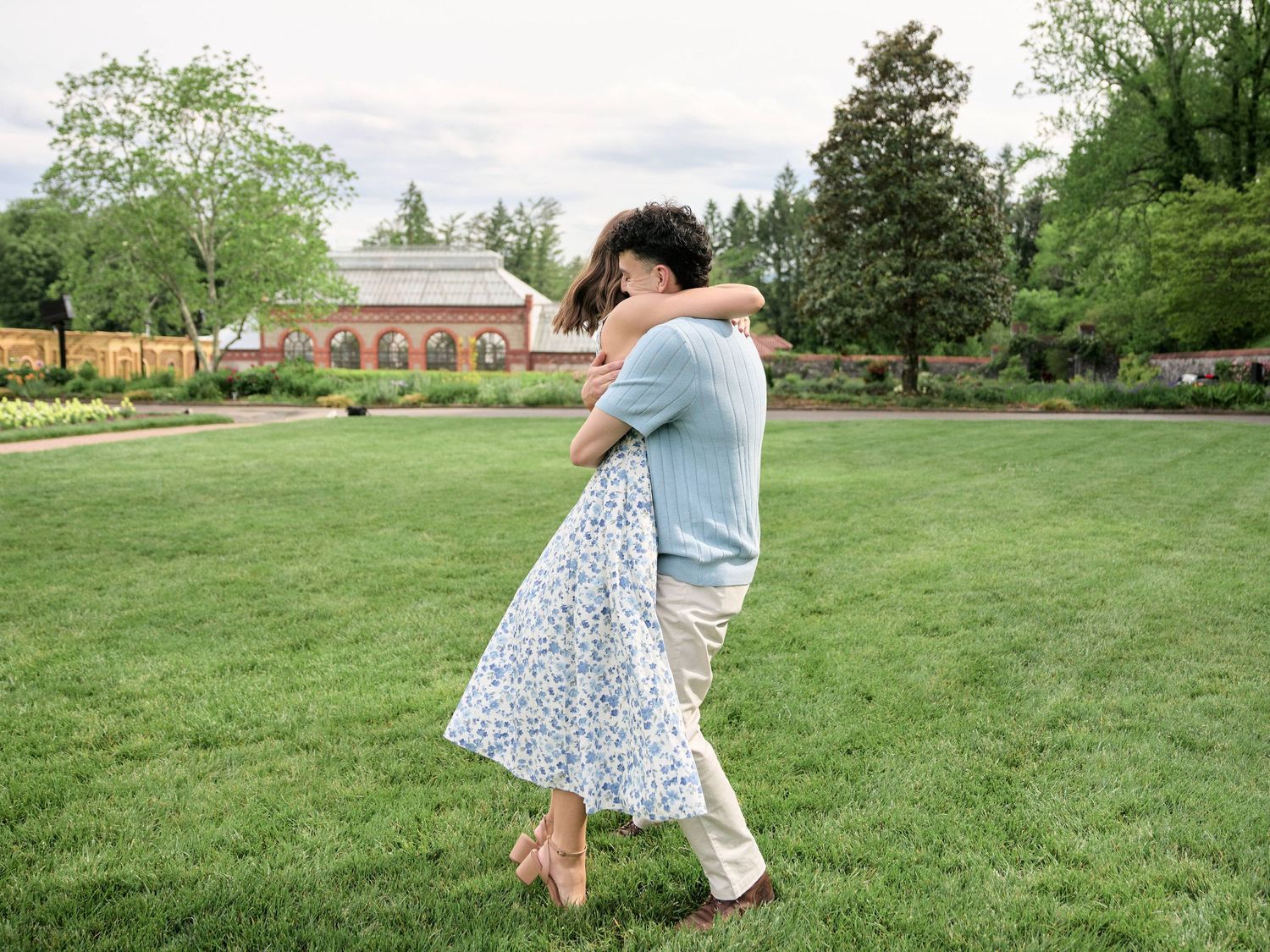 Couple embracing and dancing on lush green lawn in a romantic garden setting with trees and brick building.