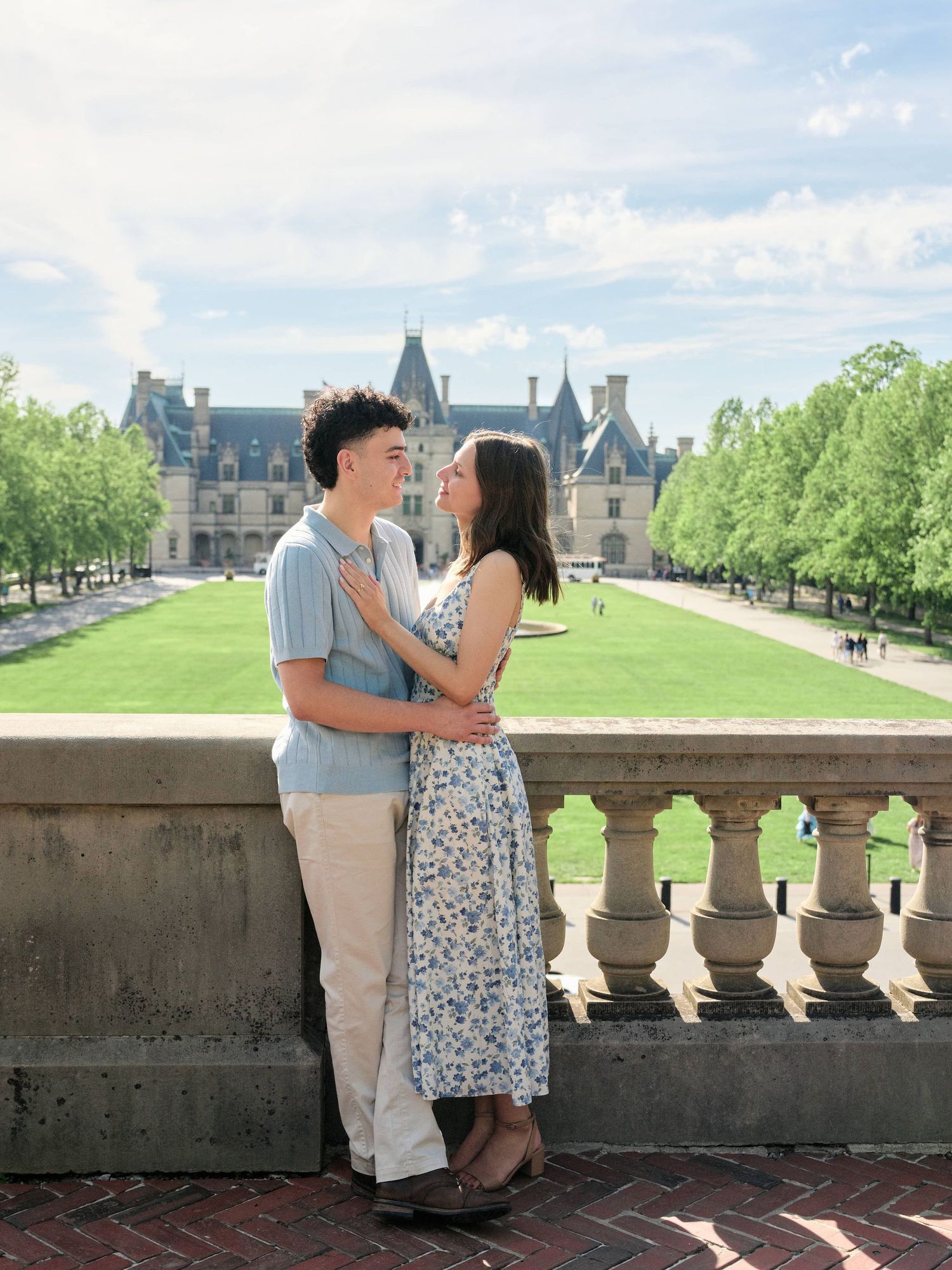 A couple embraces on a stone balcony with a grand estate and lush green lawn in the background.
