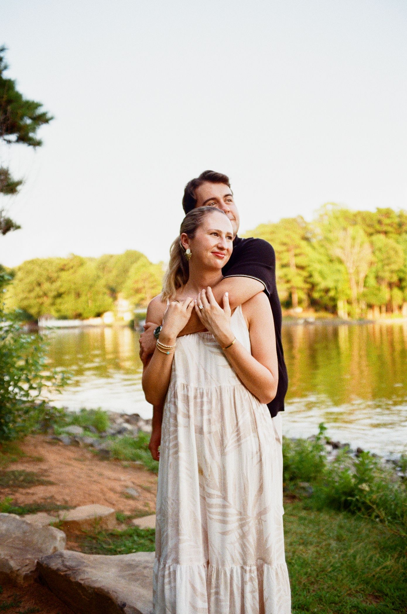 A couple embraces playfully by a serene lake during a summer photo session in a natural outdoor setting.