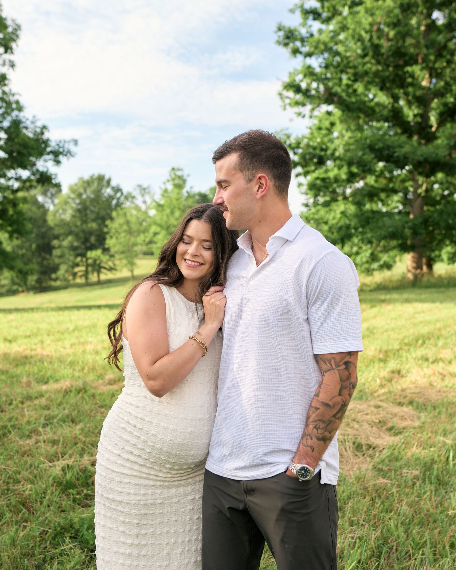 Pregnant woman in white dress embraced by tattooed partner in white polo during outdoor maternity shoot.