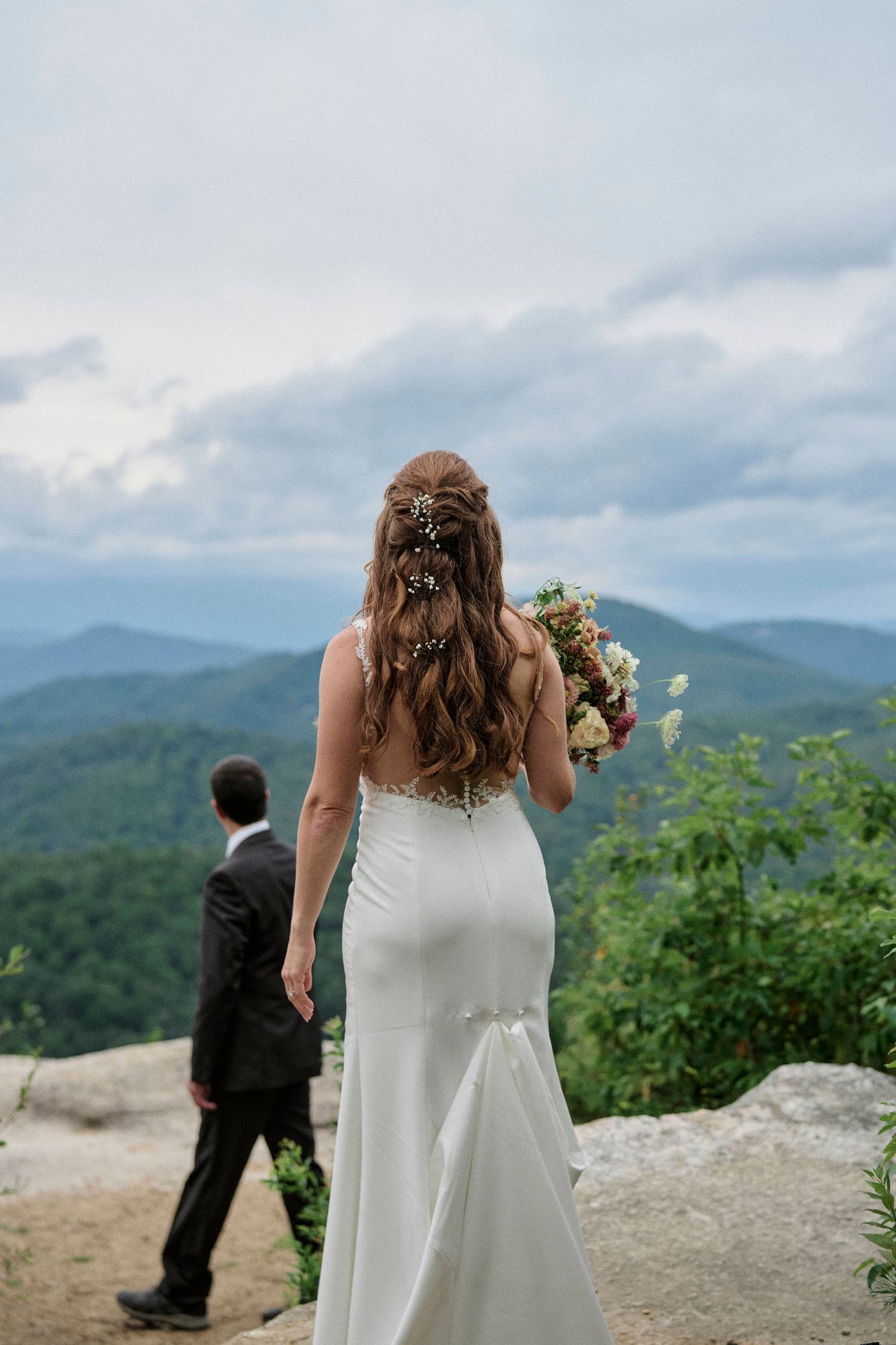 A bride in a fitted white dress holds a bouquet while standing on a mountaintop overlook with scenic mountain views.