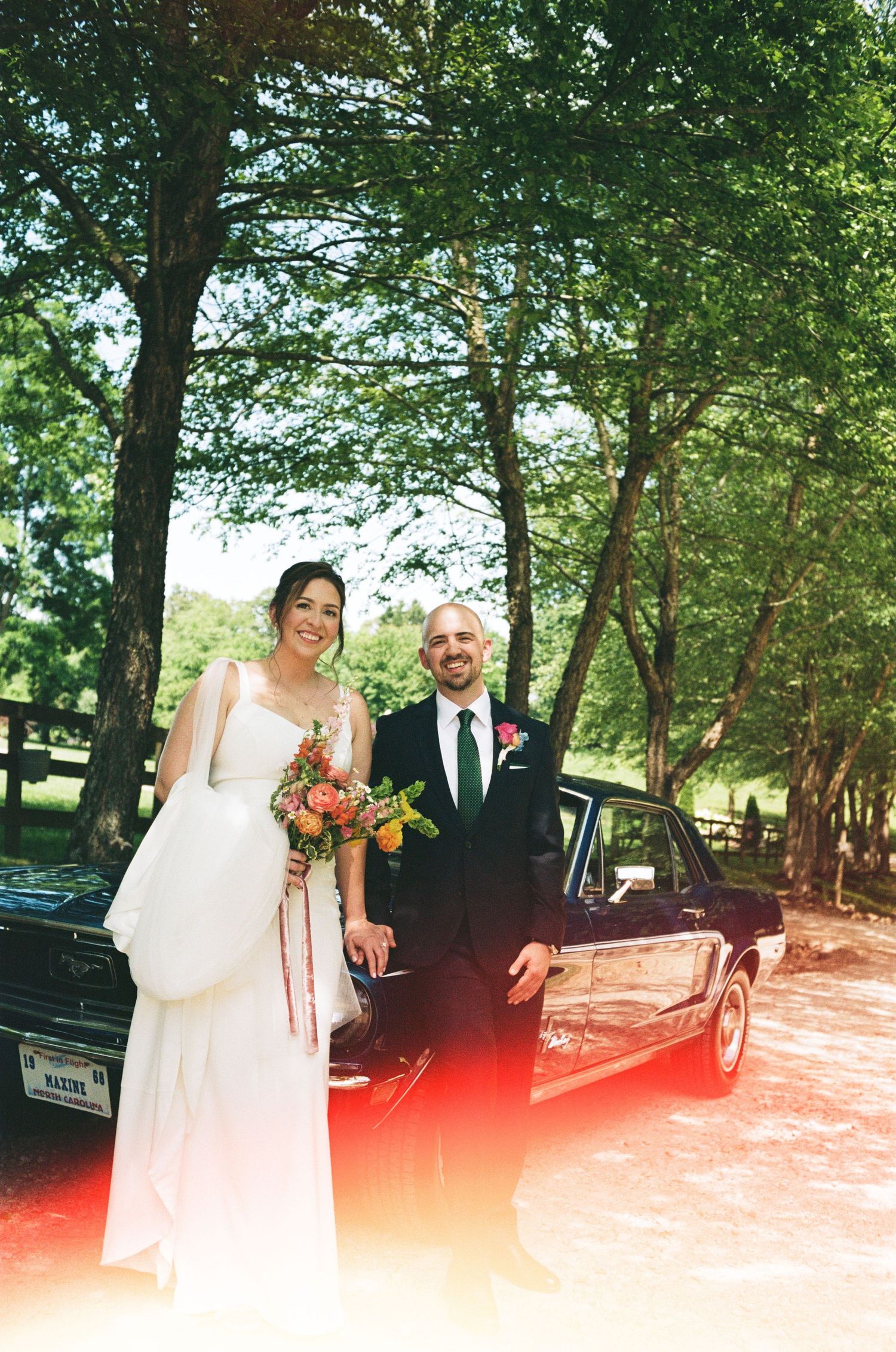A sunny outdoor wedding photo by a classic car surrounded by trees.