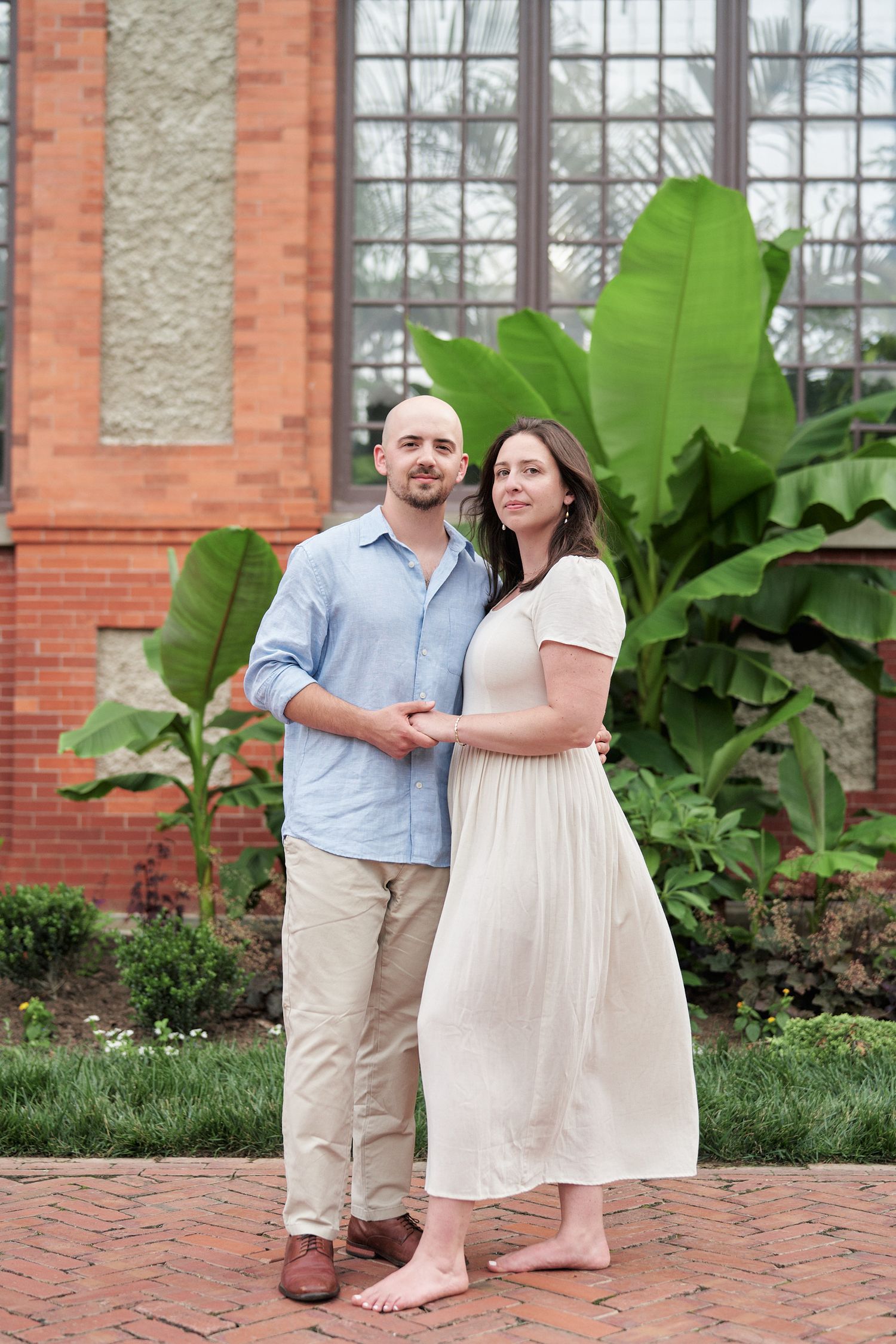 A couple poses together against a brick building with large tropical plants.