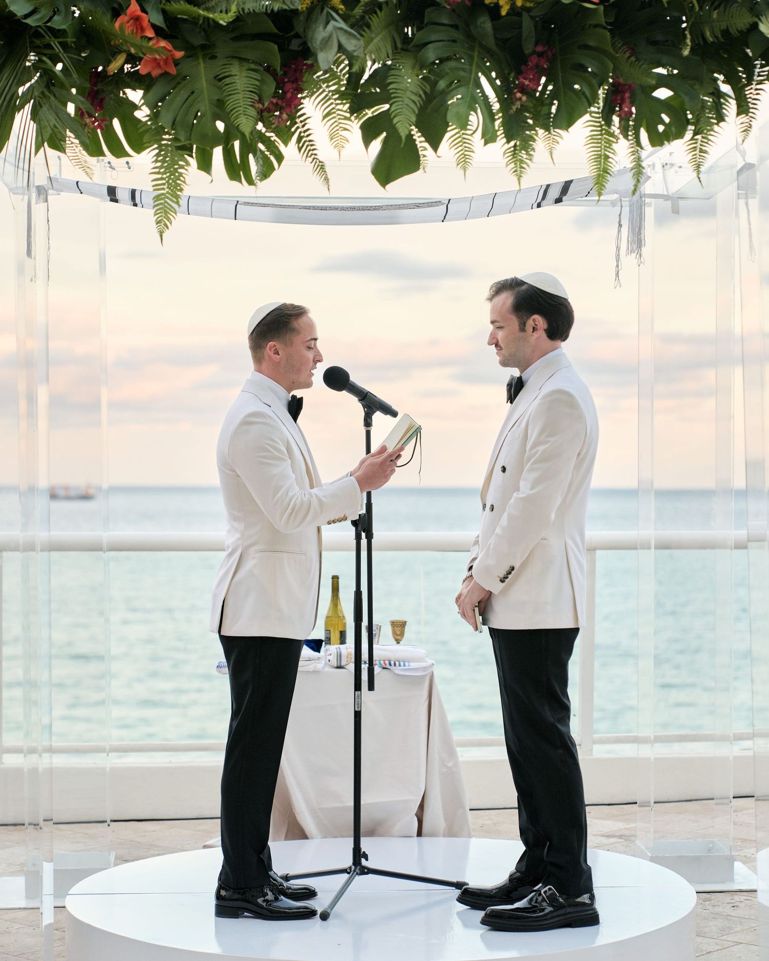 Two people in matching white tuxedo jackets stand together during a beachside ceremony under a floral-adorned canopy.