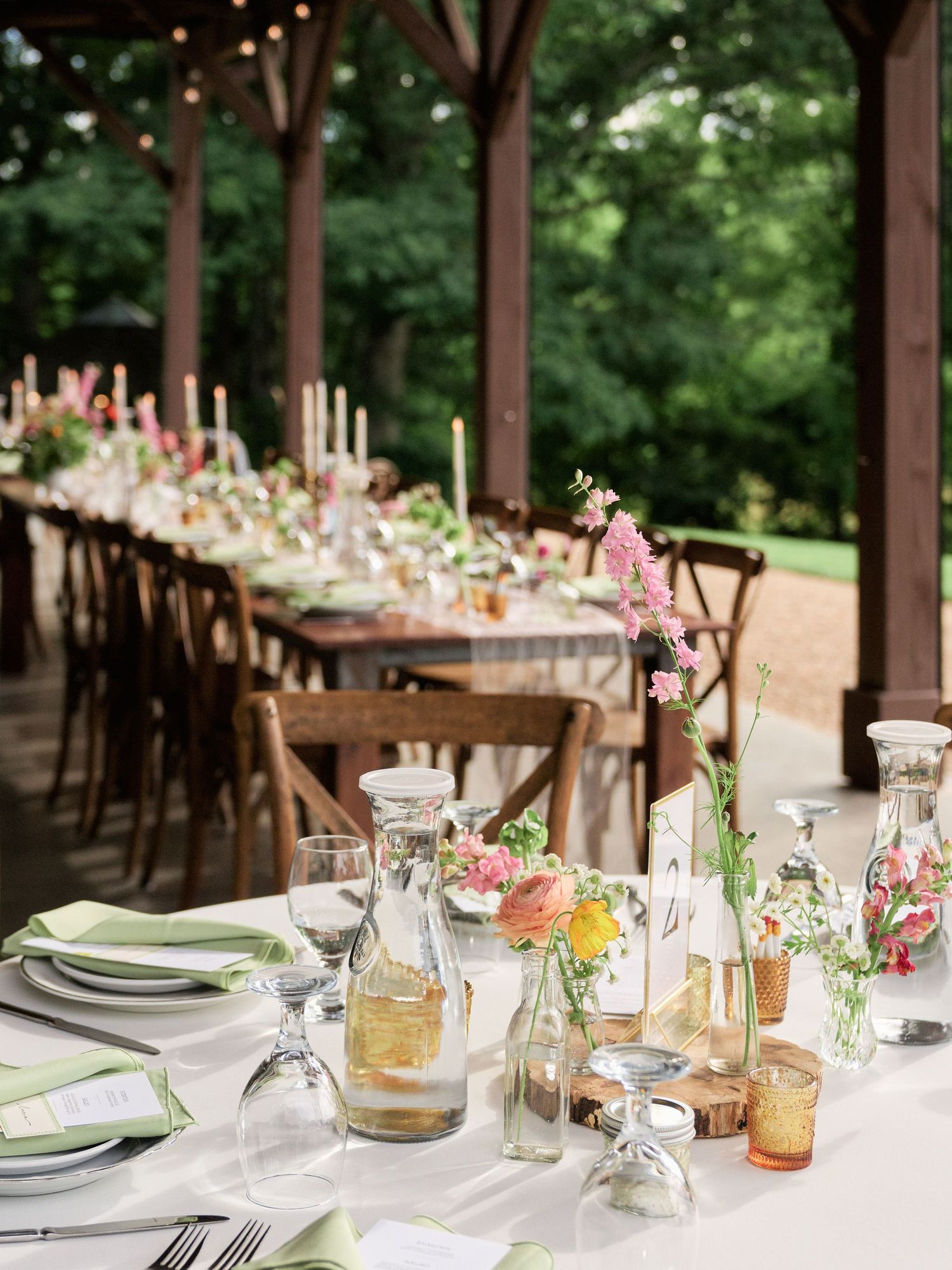 Long wooden farmhouse table set for a wedding reception under a covered pavilion with string lights.