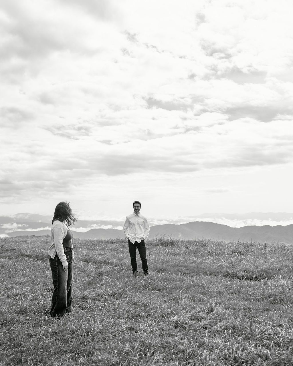 Two people stand in a black and white photograph on a grassy hilltop with mountainous terrain in the background.