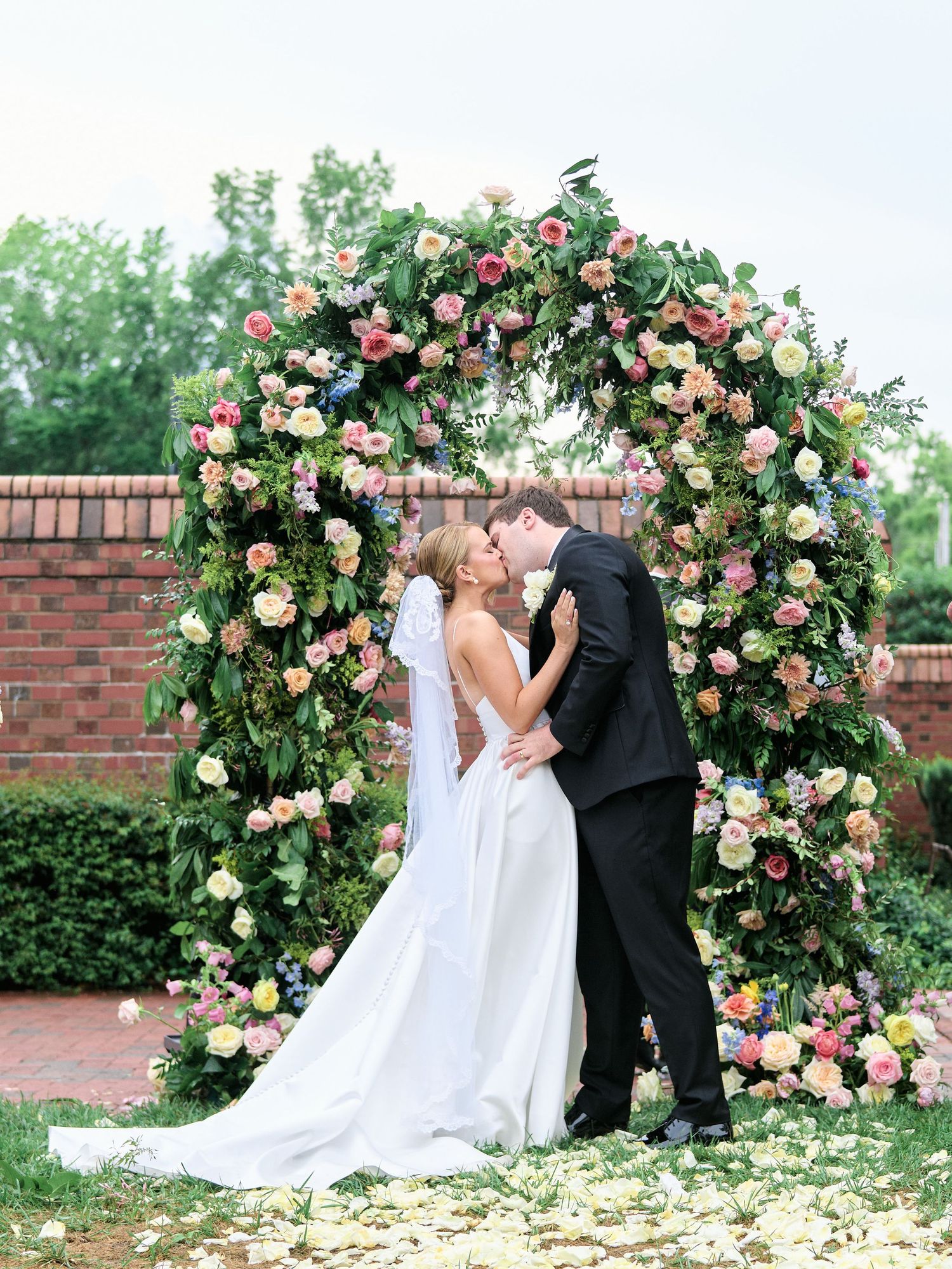 Newlyweds sharing their first kiss under an elaborate rose-covered wedding arch.