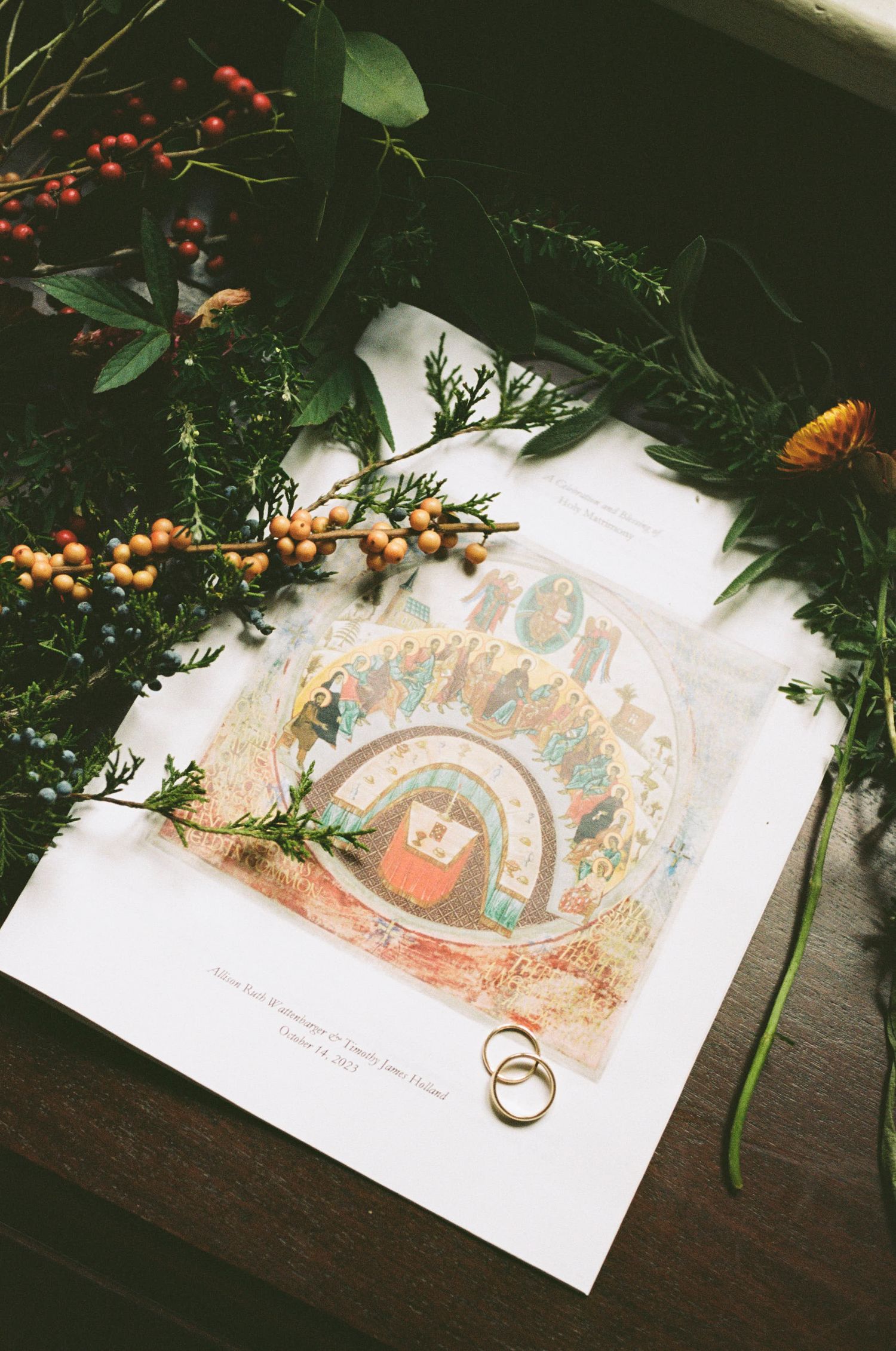 Wedding rings and floral decor lay atop an artistic illustration in a book with orange berries and greenery.