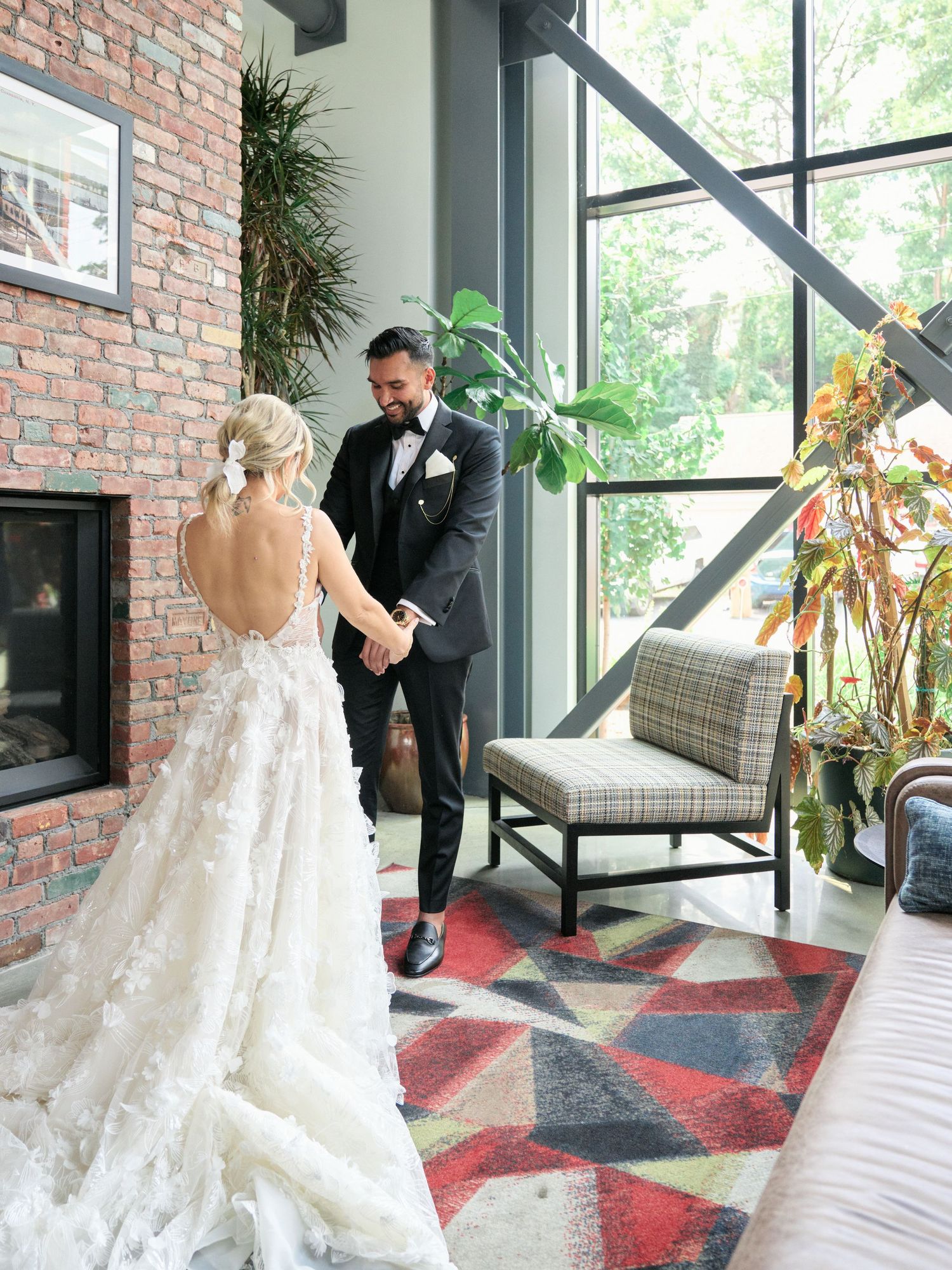 First look moment with couple in elegant wedding attire standing by brick wall and tall windows with modern decor.