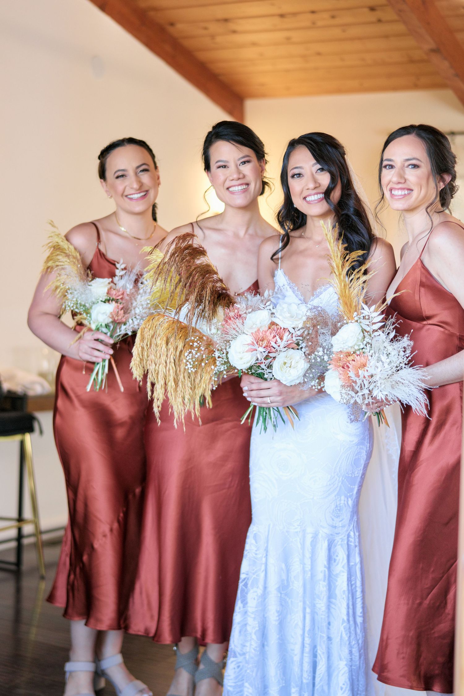 Wedding party poses together wearing rust-colored bridesmaid dresses and holding dried floral bouquets.