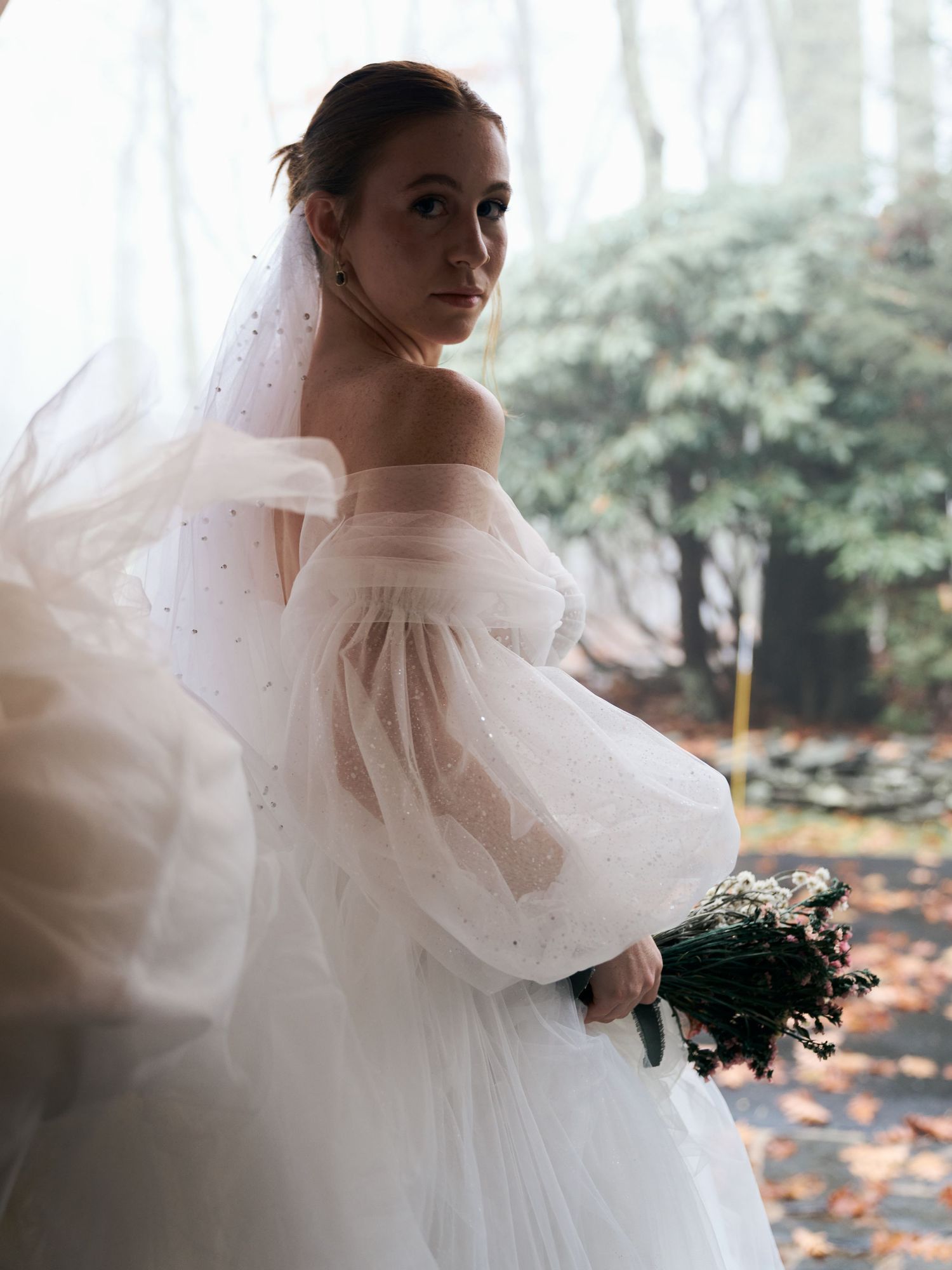 Elegant bride in off-shoulder wedding dress with puffy sleeves holding bouquet in outdoor setting.
