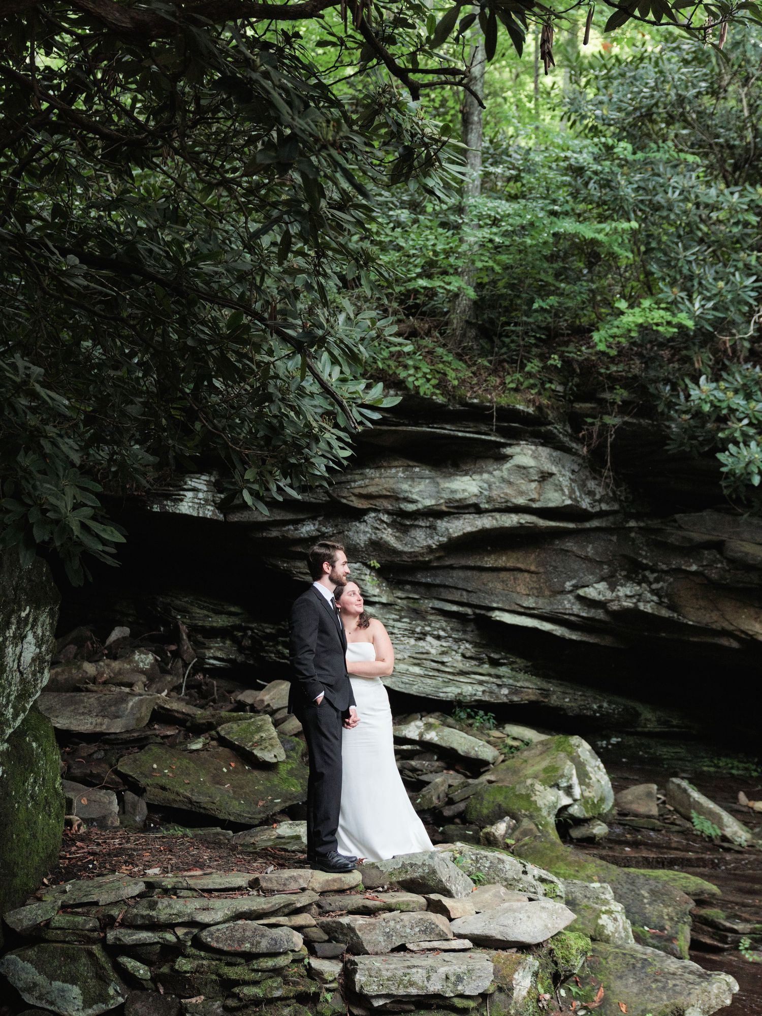 A couple poses together on mossy rocks against a backdrop of natural stone walls in a wooded outdoor setting.