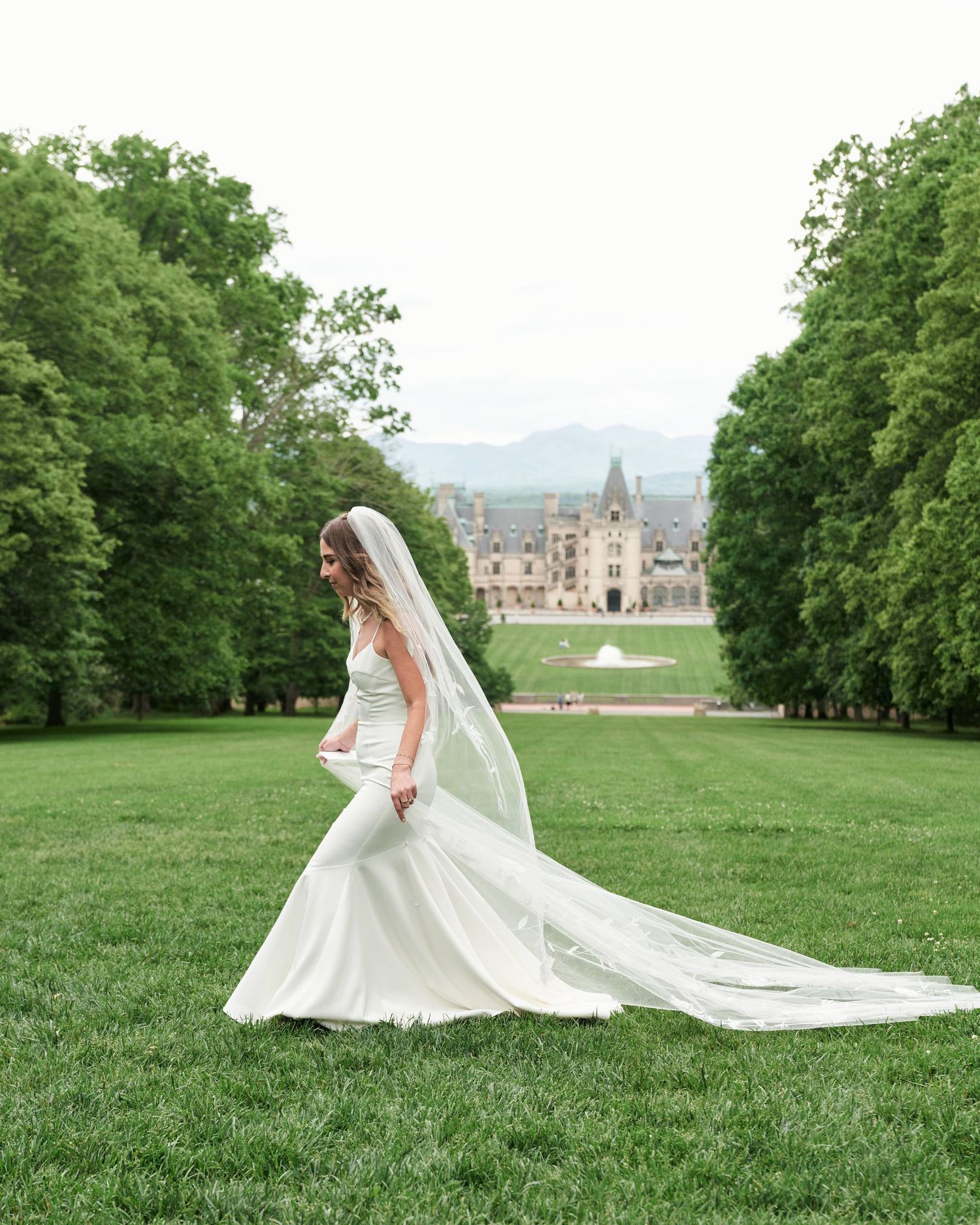 A flowing cathedral-length veil trails behind a bride in a white gown as she poses on a grassy hillside near a castle.
