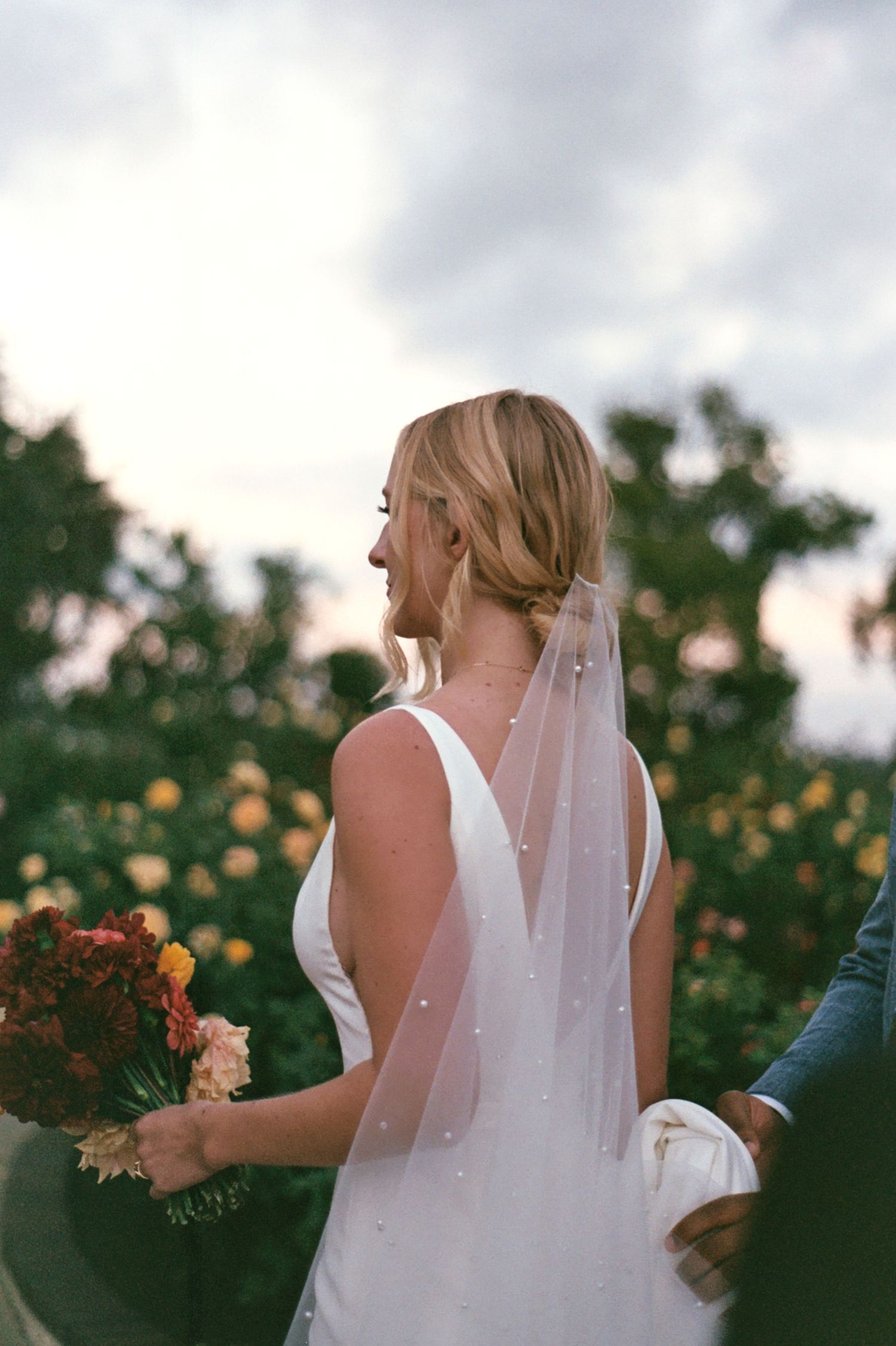 A bride in a sleeveless white gown and veil stands in a garden with golden sunlight filtering through trees at sunset.