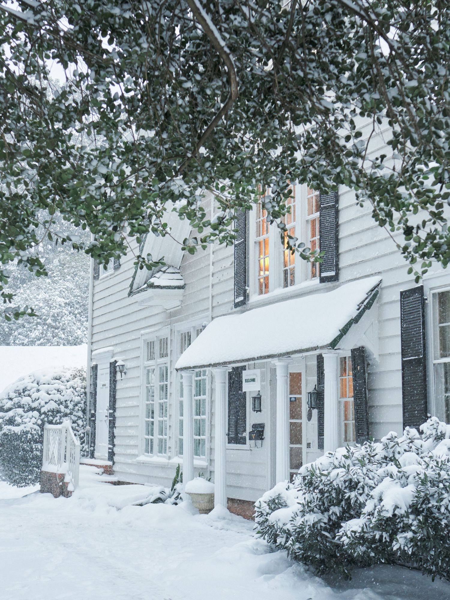 Snow-covered white colonial home with dark shutters, warm glowing windows, and festive holiday lights during a winter snowstorm.
