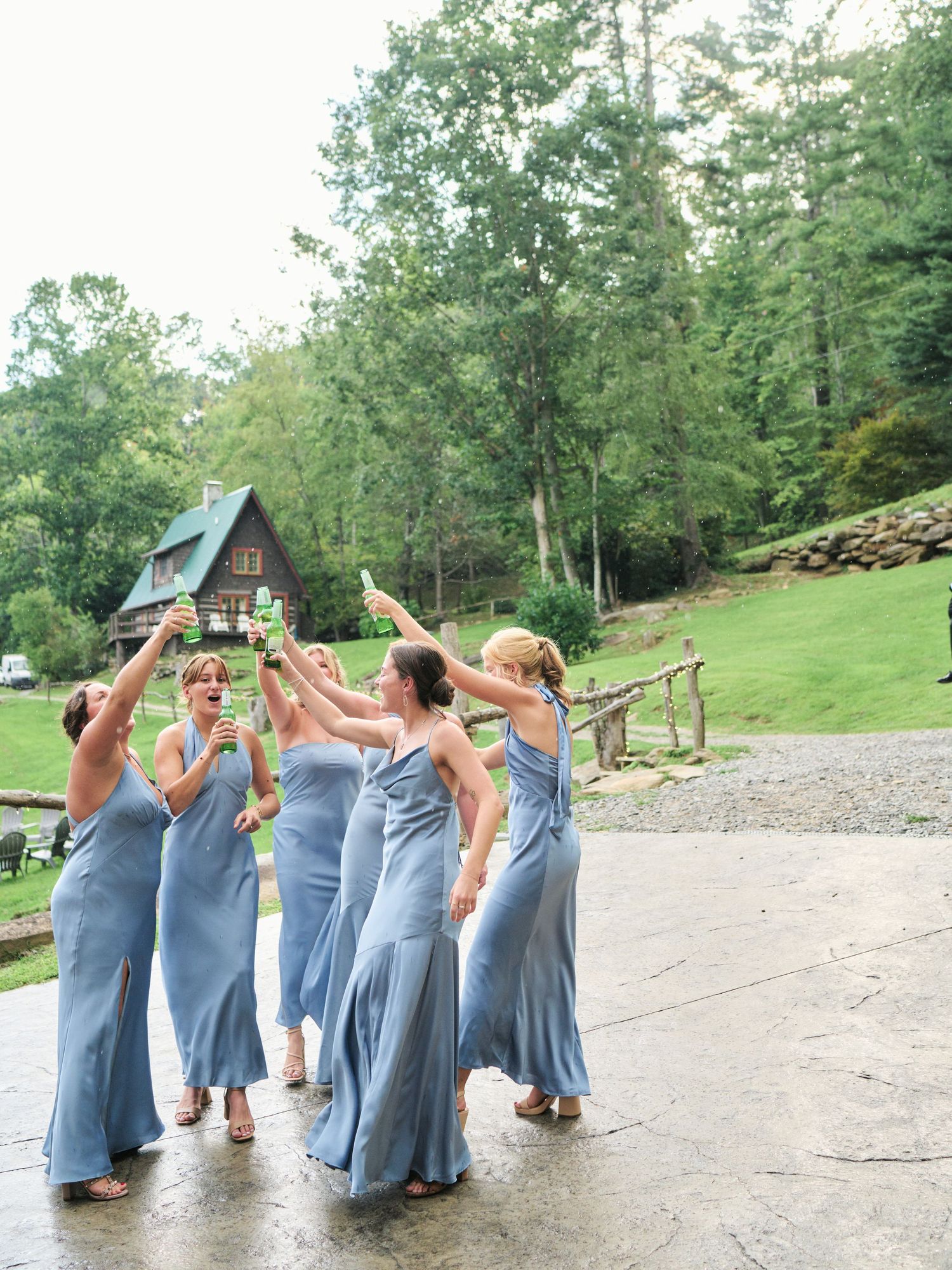 Bridesmaids in blue gowns toast with champagne outdoors near a rustic cabin surrounded by lush green trees.