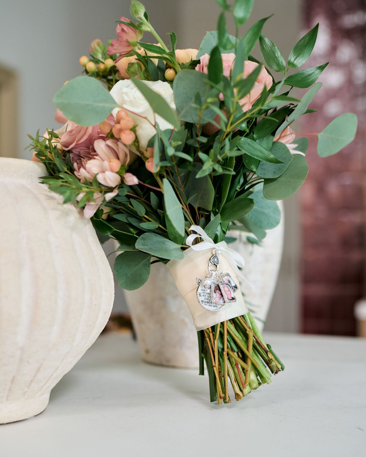 Elegant wedding bouquet with pink roses, eucalyptus, and a photo charm tied with ribbon, displayed on a white surface.