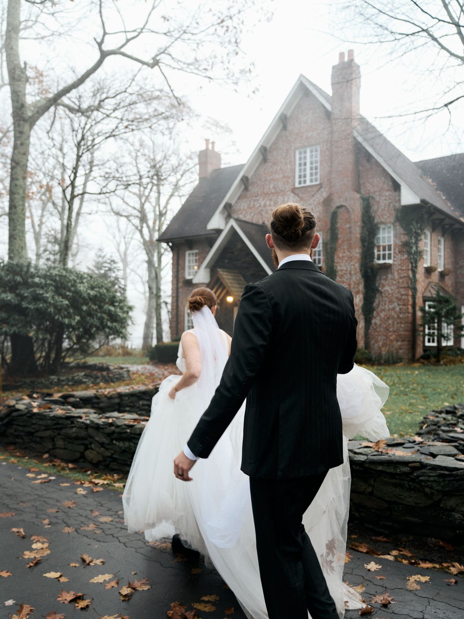 A bride in white and groom in black walk along a misty path towards a brick Gothic-style house with autumn leaves.