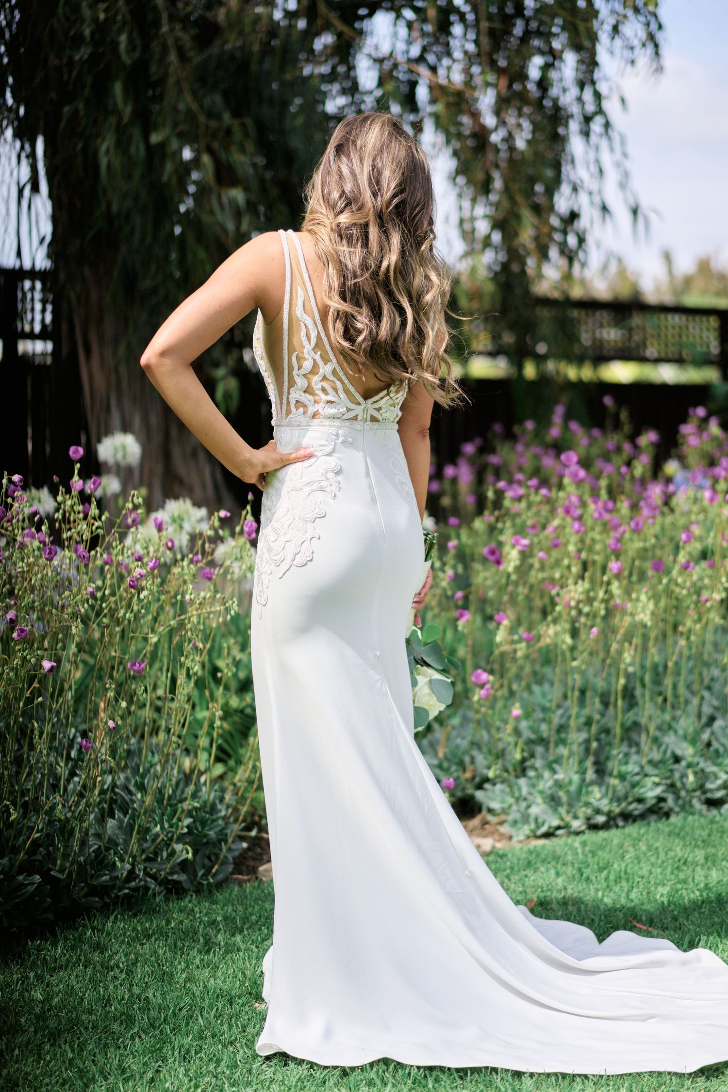 Elegant white wedding dress with lace detail shown in a garden with purple wildflowers.
