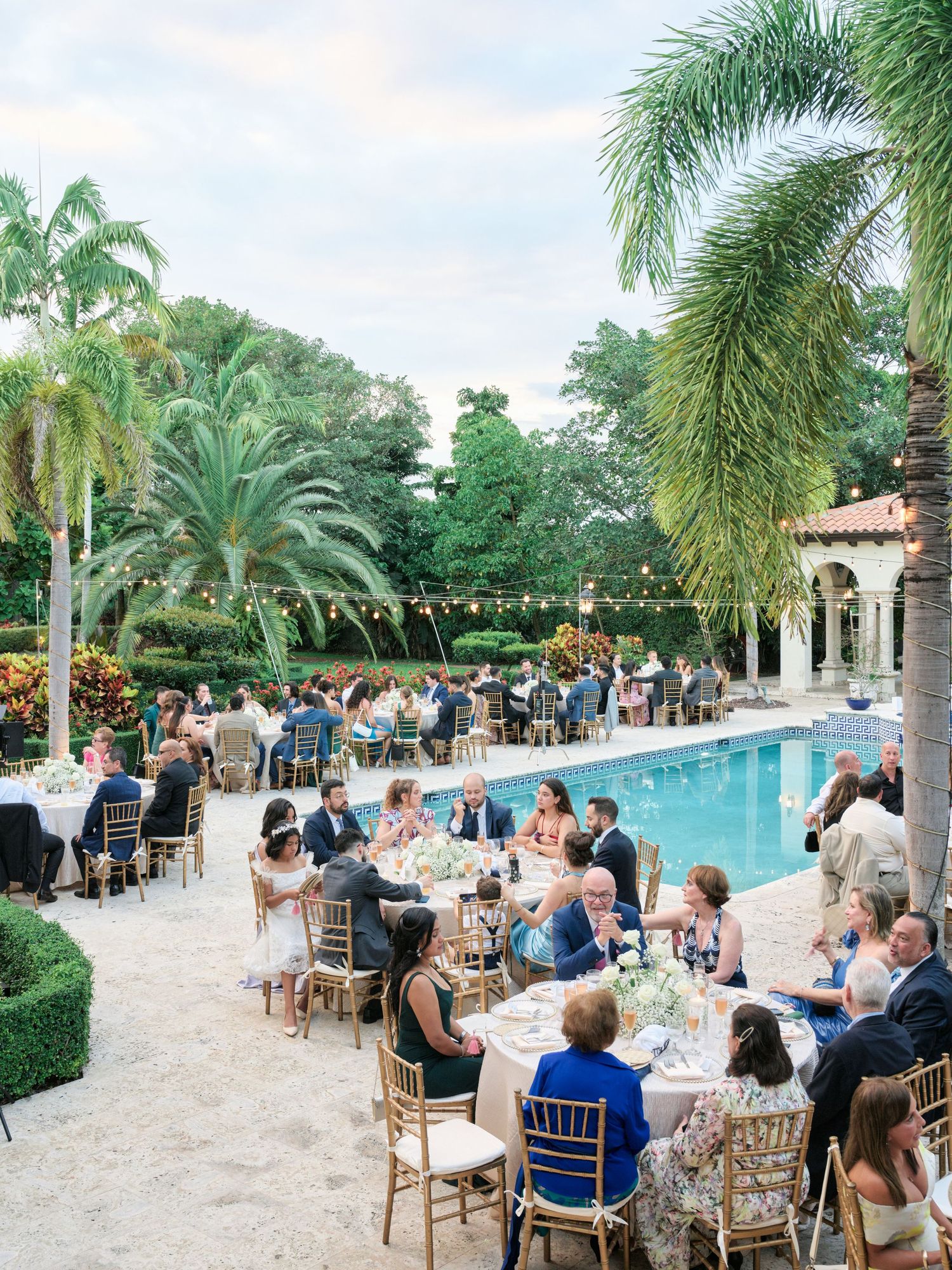 Outdoor wedding reception by a pool surrounded by palm trees with string lights and dining tables.