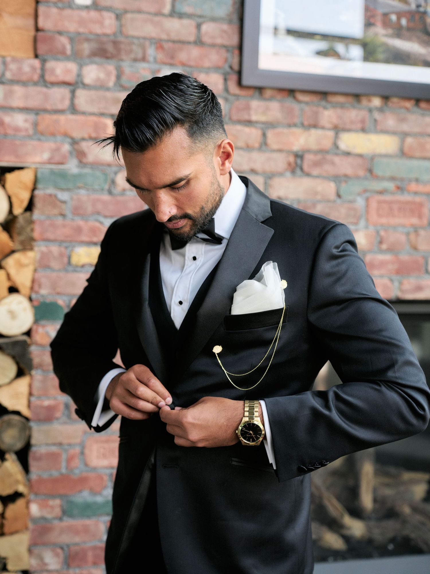 A groom in a classic black tuxedo with bow tie and pocket square adjusts his jacket against an exposed brick wall.