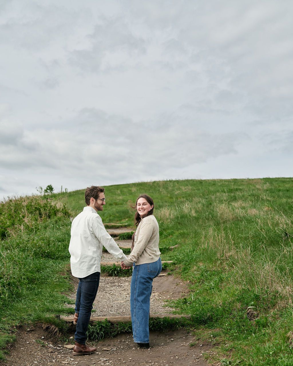 Two people holding hands on a dirt path leading up a grassy hillside under cloudy skies.