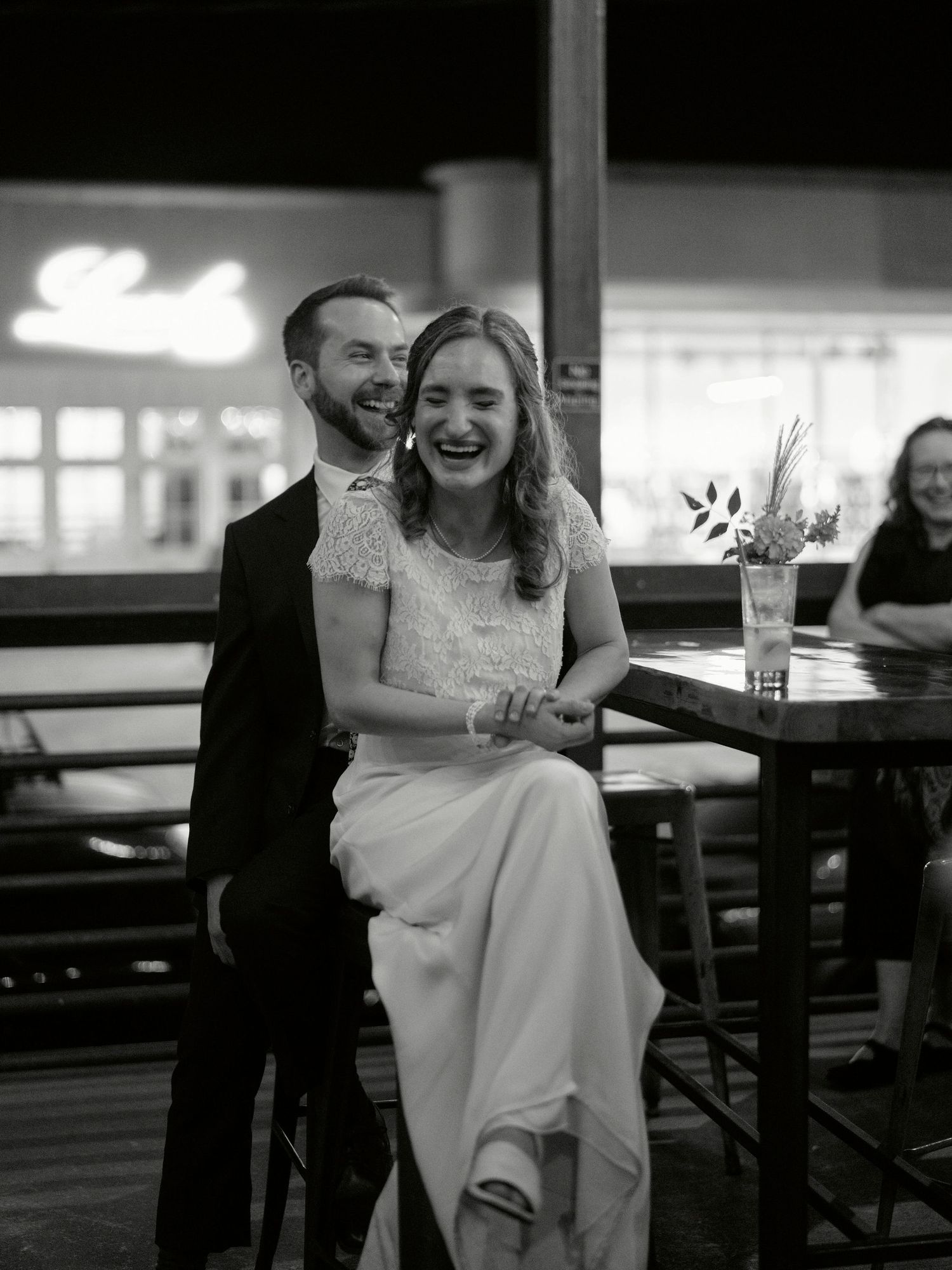 A couple shares a candid laugh while sitting at a bar during their wedding celebration.