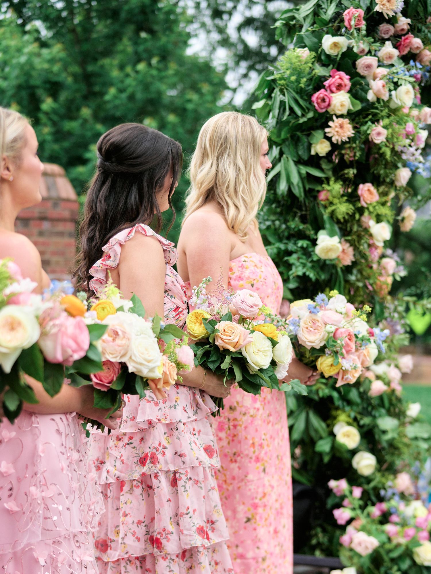 Bridesmaids in pink floral dresses holding bouquets next to a decorative flower arch.