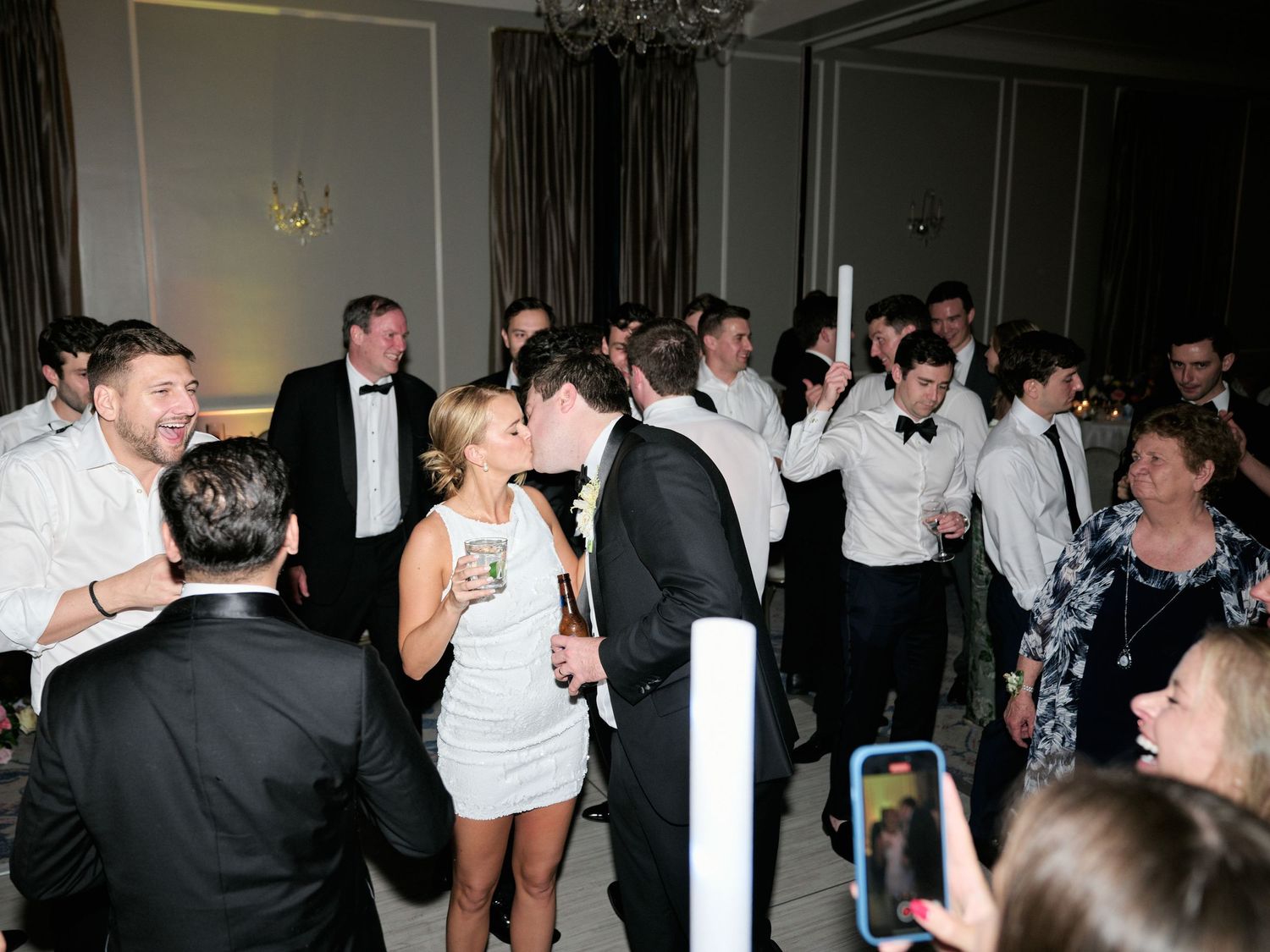 Wedding reception guests celebrate and dance in formal attire while someone takes a photo on their phone.