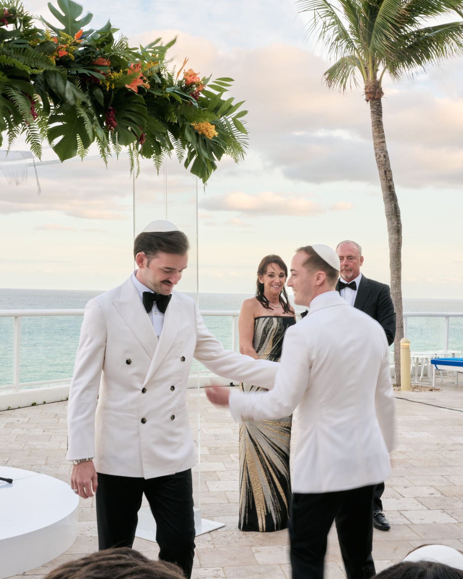A beachside ceremony takes place under a tropical floral arch with palm trees and ocean views in the background.