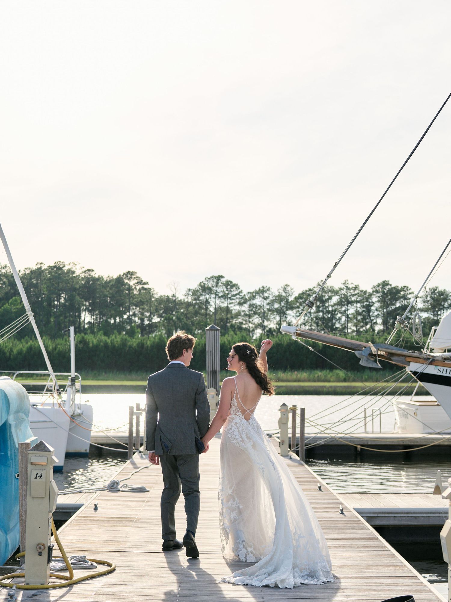 Couple in wedding attire standing on a wooden dock with sailboats and a marina in the background at sunset.