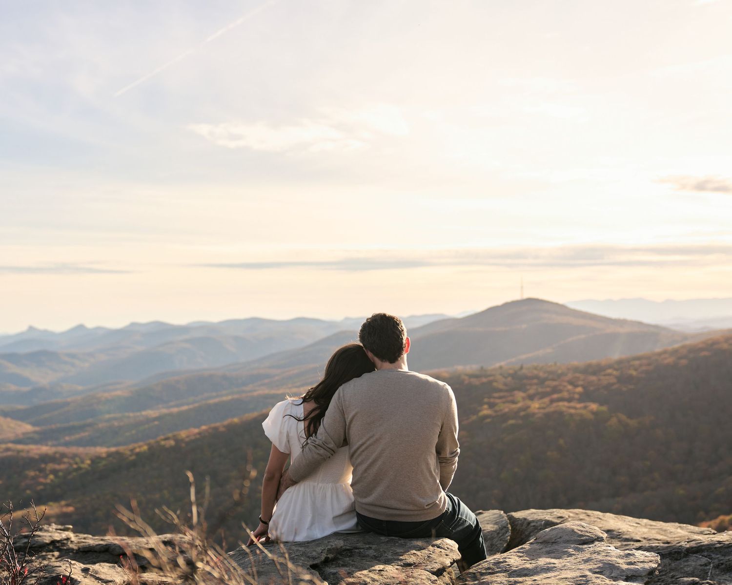 Engaged couple sit together on a mountain ledge overlooking a scenic valley during sunset in the Blue Ridge mountains.