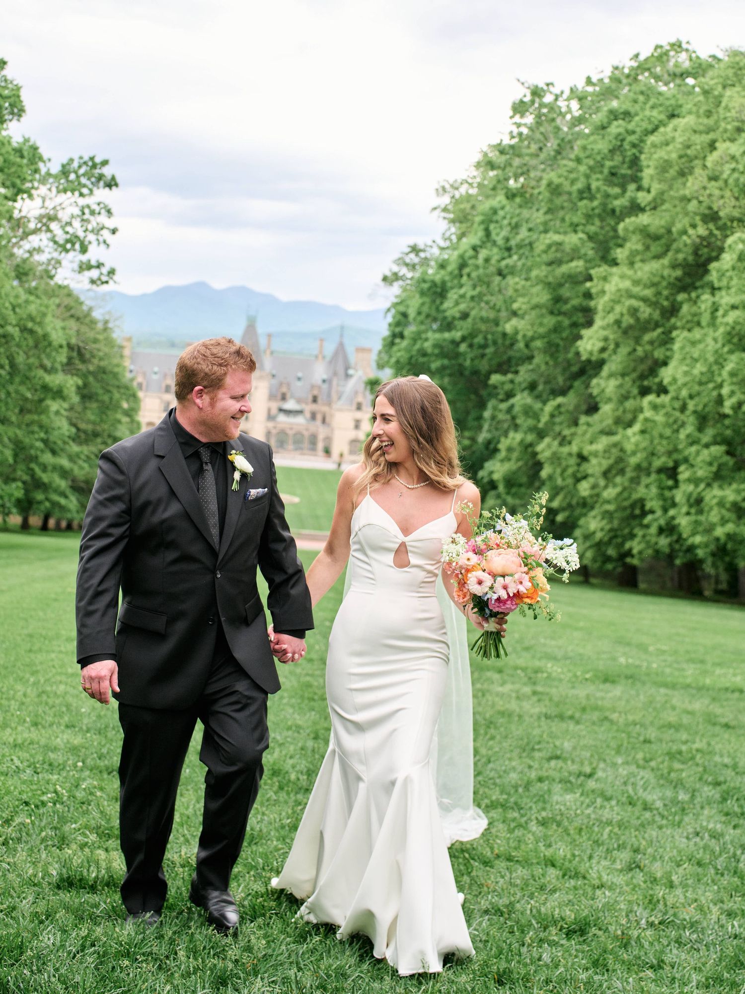 A couple in wedding attire walk together on a lush green hillside with trees and mountains in the background.