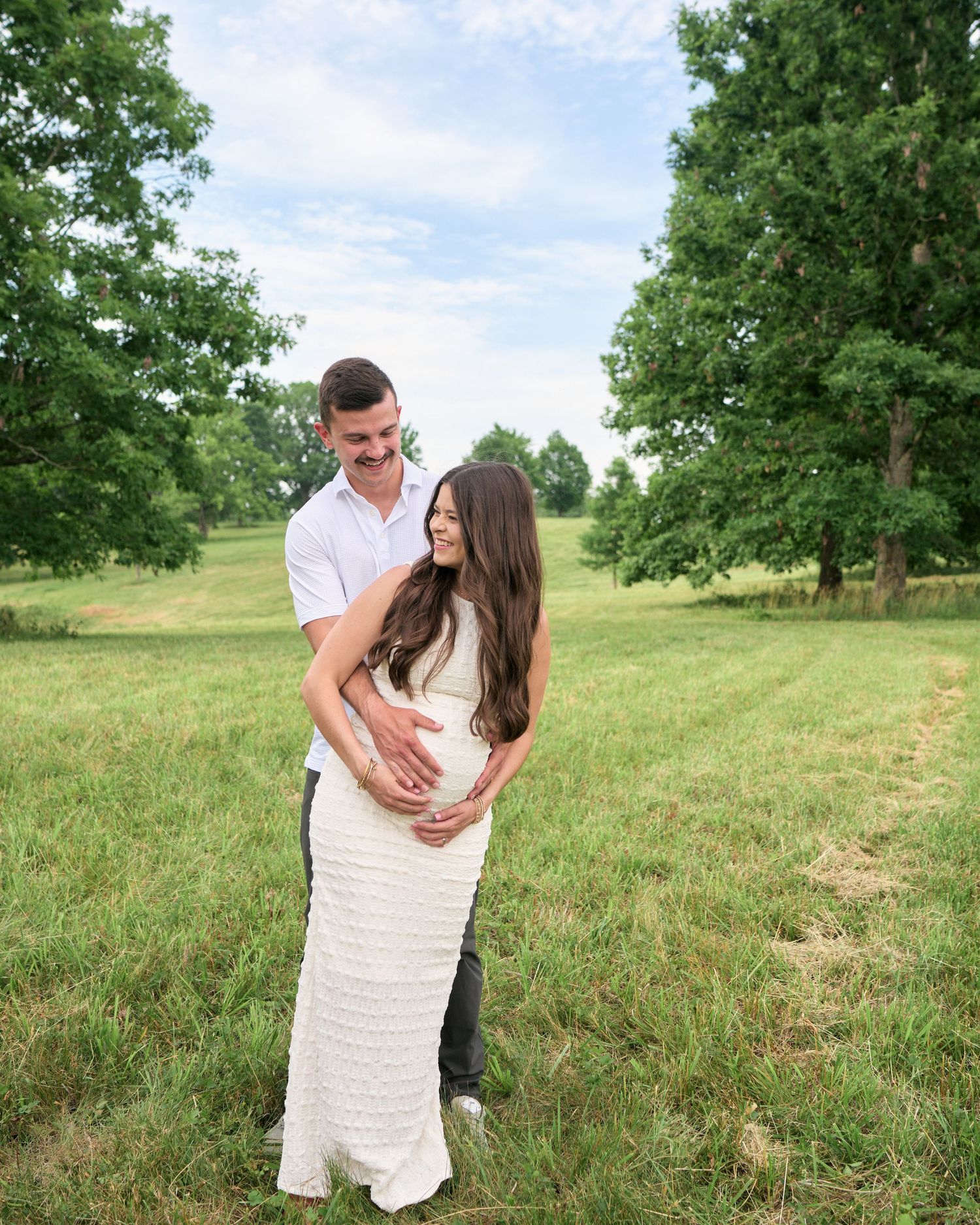 A couple poses in a green field, the man embracing the pregnant woman in a white gown, surrounded by lush trees.