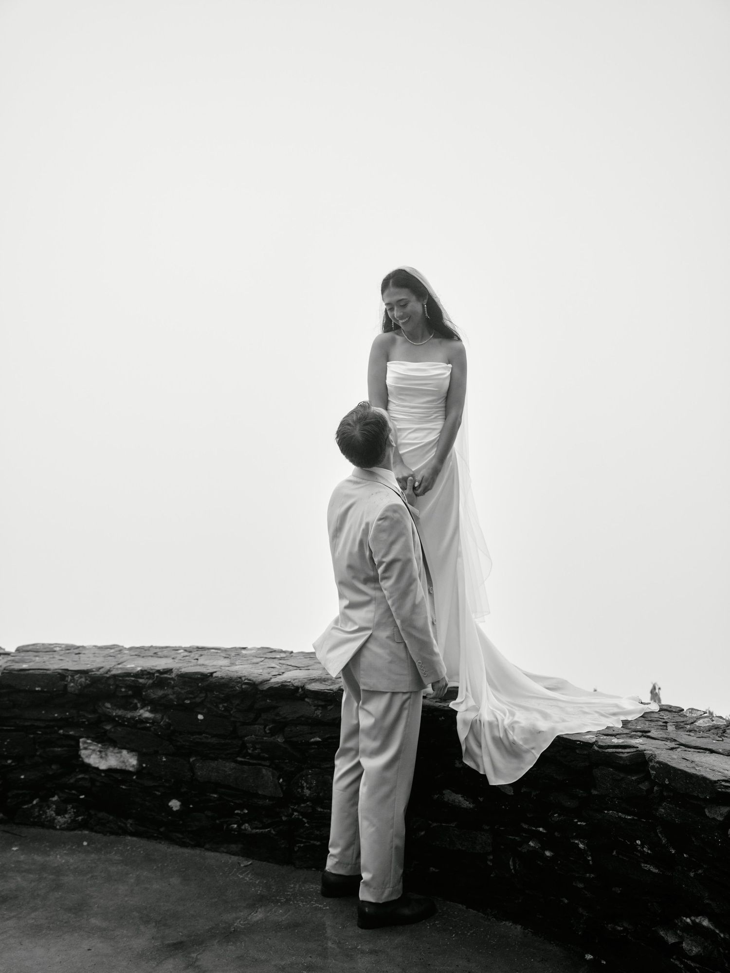 Black and white photo of a bride in a white gown and veil standing on a stone wall with a groom below her.